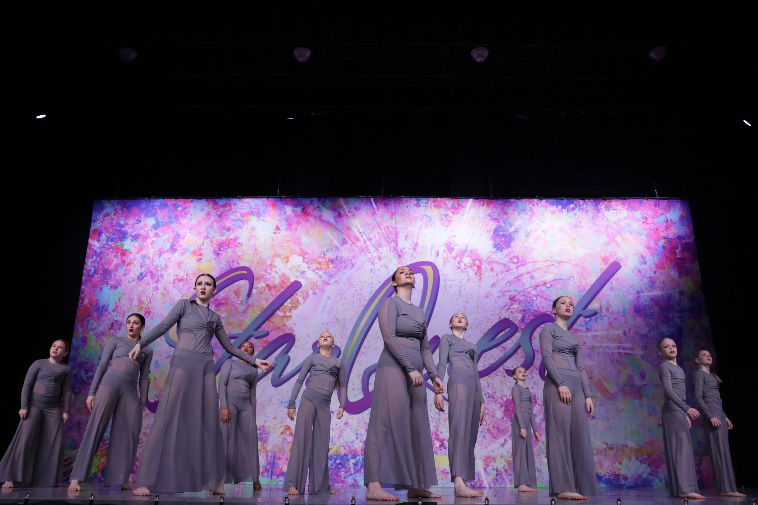 Group of young female dancers in matching gray costumes performing on stage with a colorful, abstract backdrop.