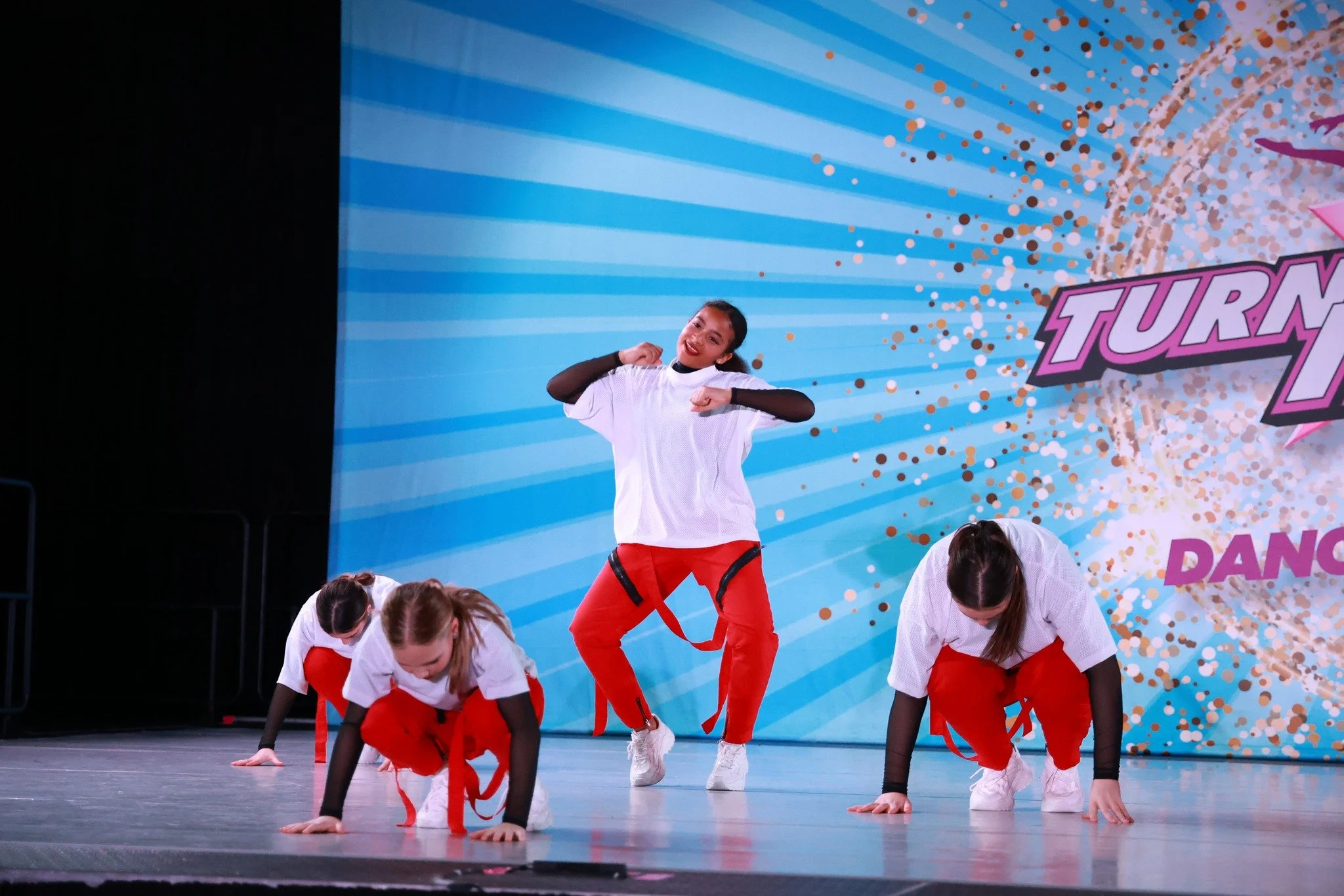 Group of young women performing a dance routine on stage, with the lead dancer in the center. They are dressed in white T-shirts, black arm sleeves, red pants, and white shoes. The stage background has a blue radial pattern with gold and pink confett