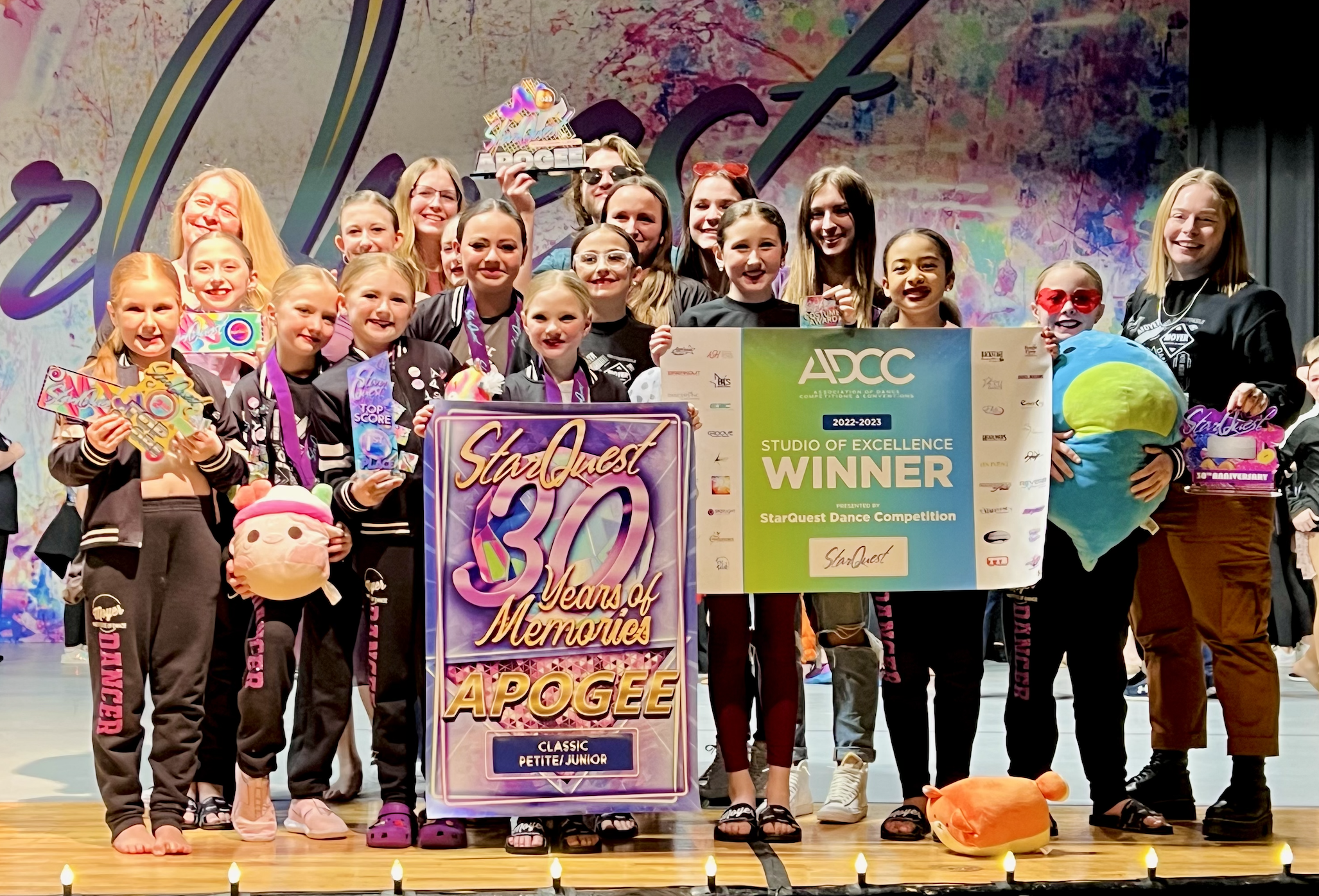 Group of young dancers on stage celebrating winning the StarQuest dance competition, holding awards, medals, and plush toys, with a large banner and stage backdrop.