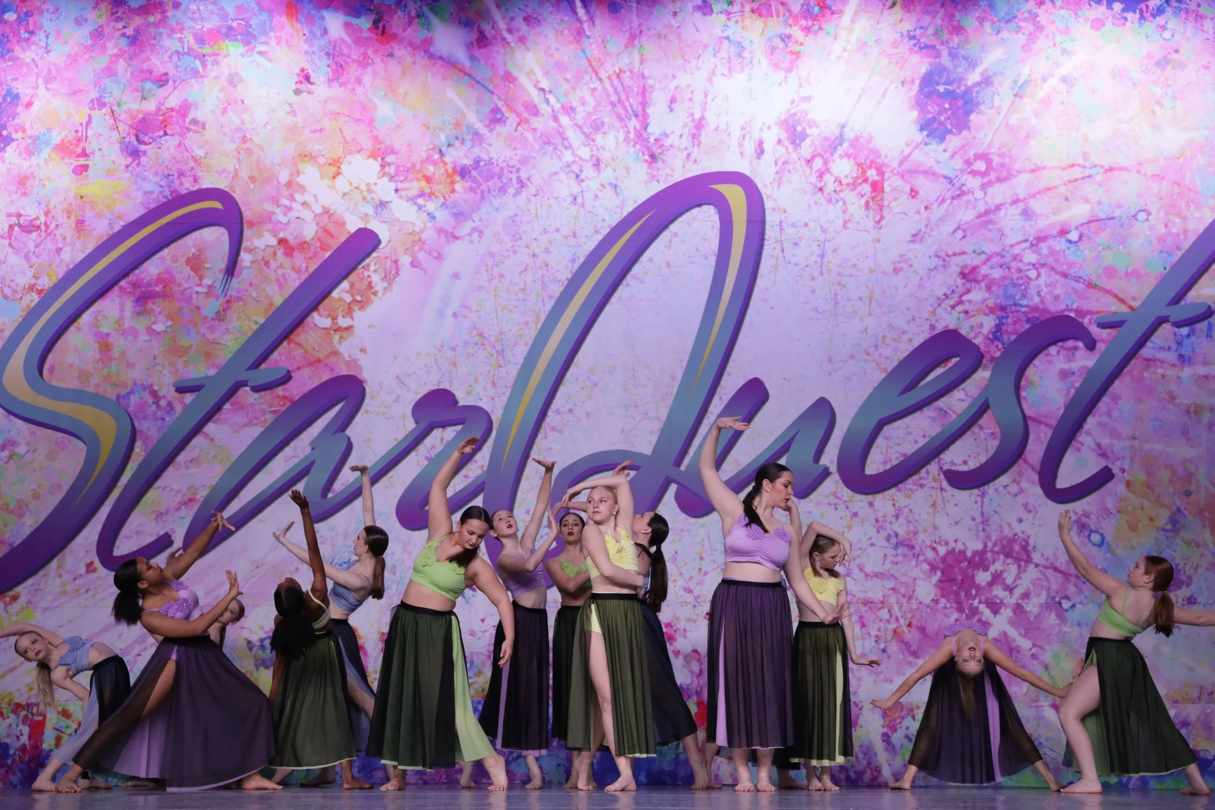 Group of young female dancers performing on stage in front of a backdrop with the word 'StarQuest' and colorful splashes of pink, purple, and yellow paint.