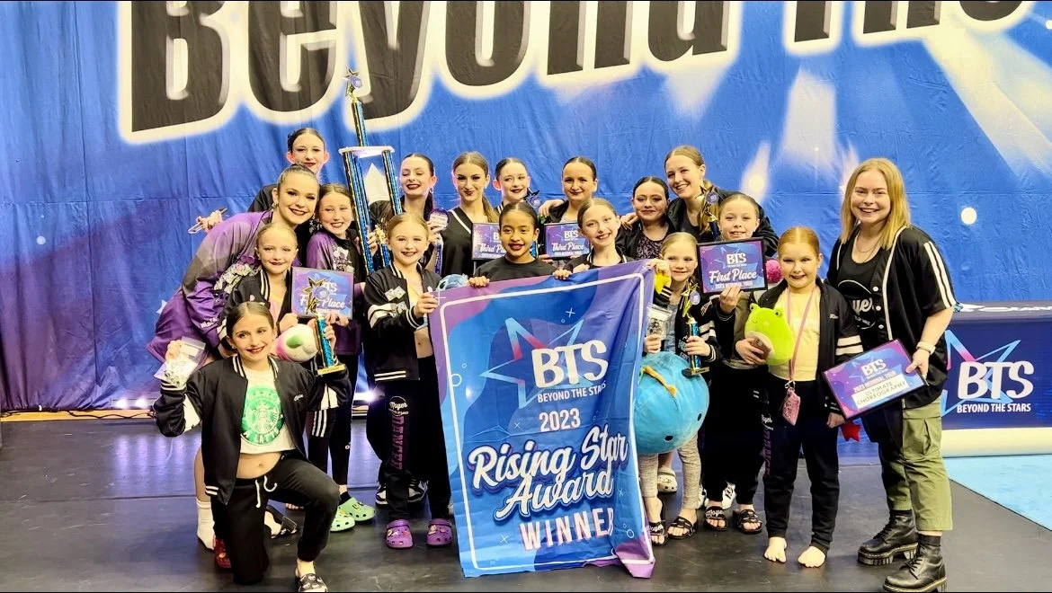 A group of young girls and their coach posing on stage at a cheerleading competition, holding awards and trophies, with a large BTS Rising Star Award banner.