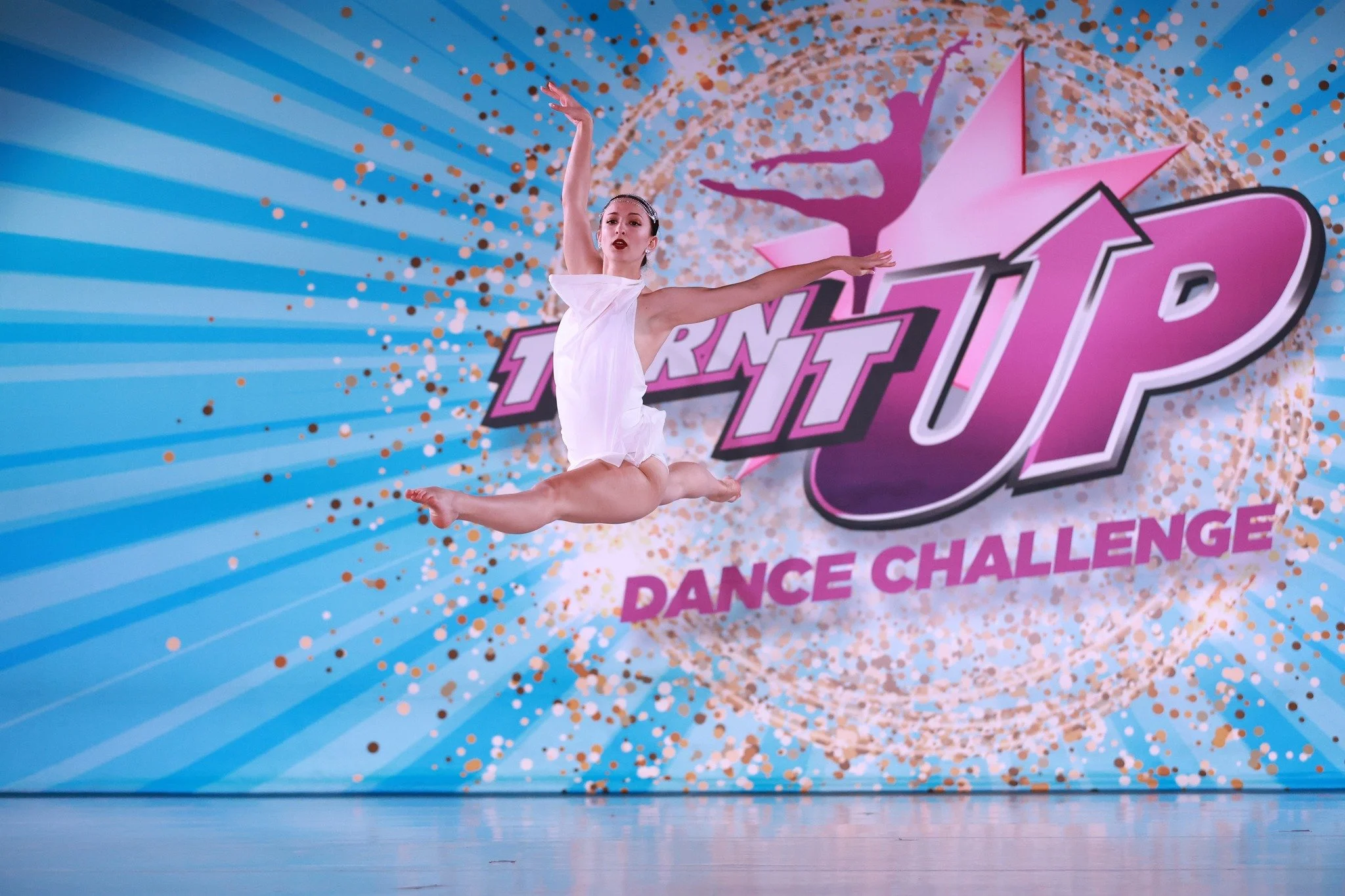 A dancer in a white dress is performing a leap on stage with a backdrop that reads 'TURN IT UP DANCE CHALLENGE' in pink and purple font, with a star and confetti design.