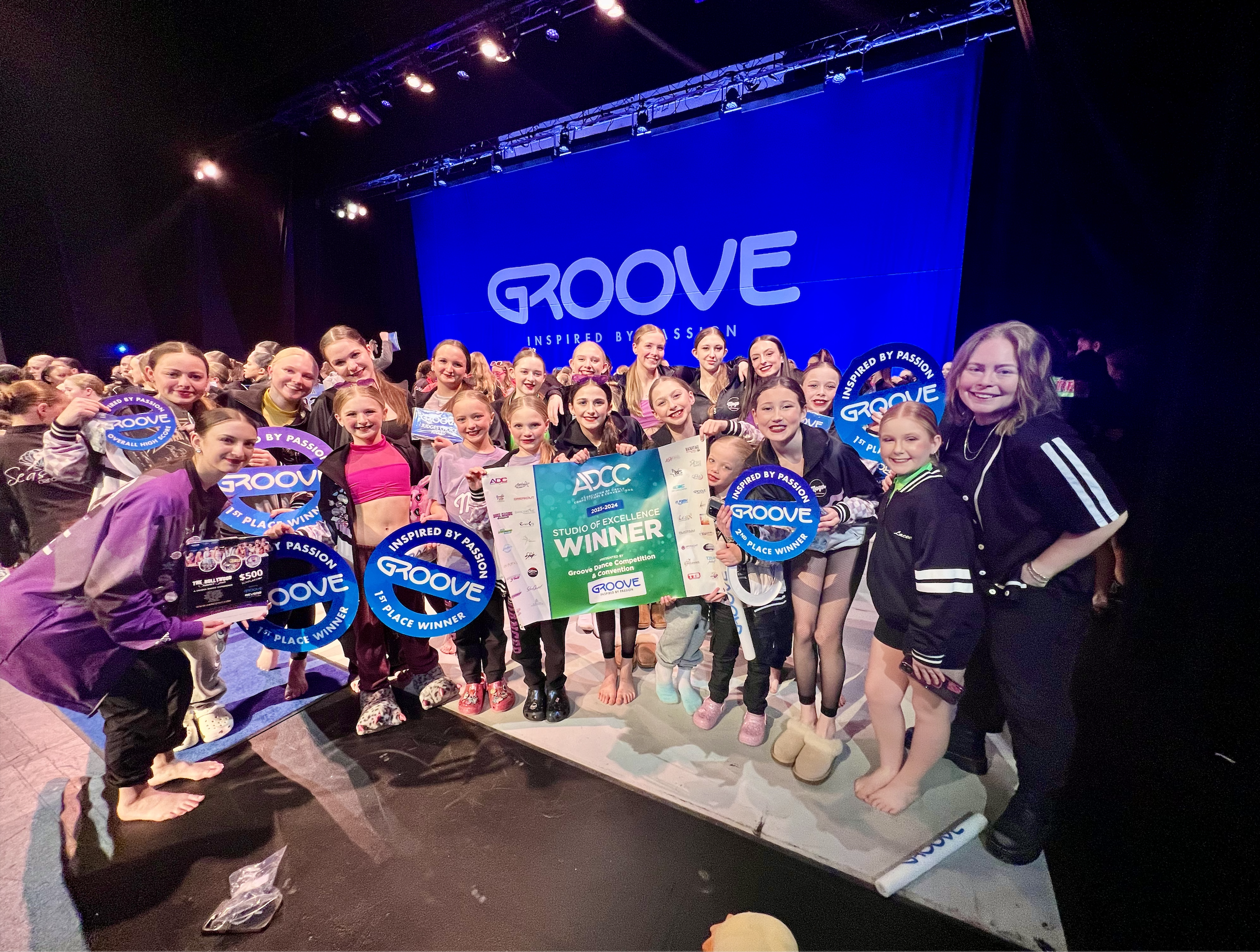Group of young dancers and instructor celebrating after a dance competition, holding trophies and a large certificate, on stage with a large blue screen displaying the word 'GROOVE' and the phrase 'Inspired by Passion'.