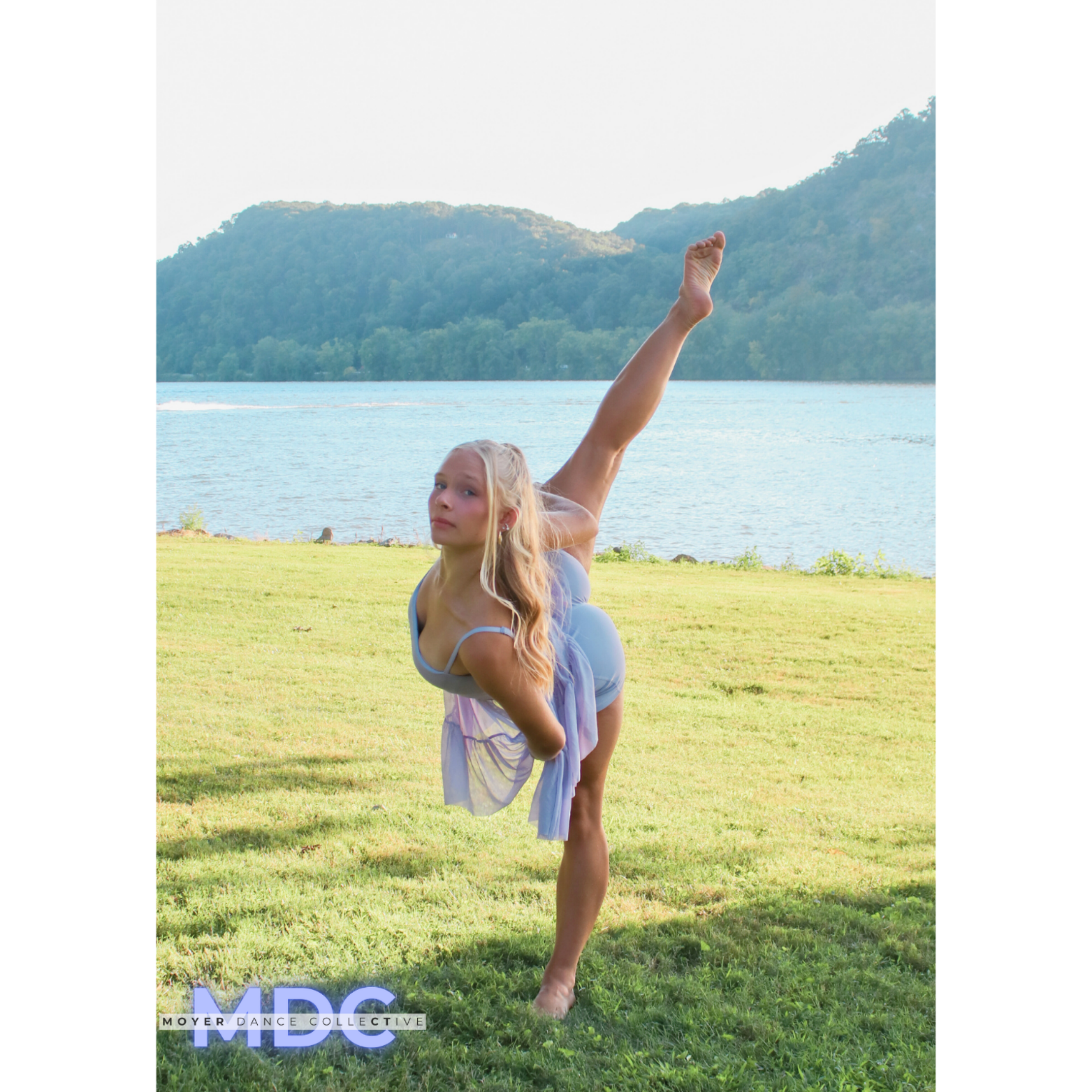 A young woman with long blonde hair practicing yoga outdoors by a lake, performing a standing one-legged bend with arms behind her back, in a scenic setting with mountains in the background.