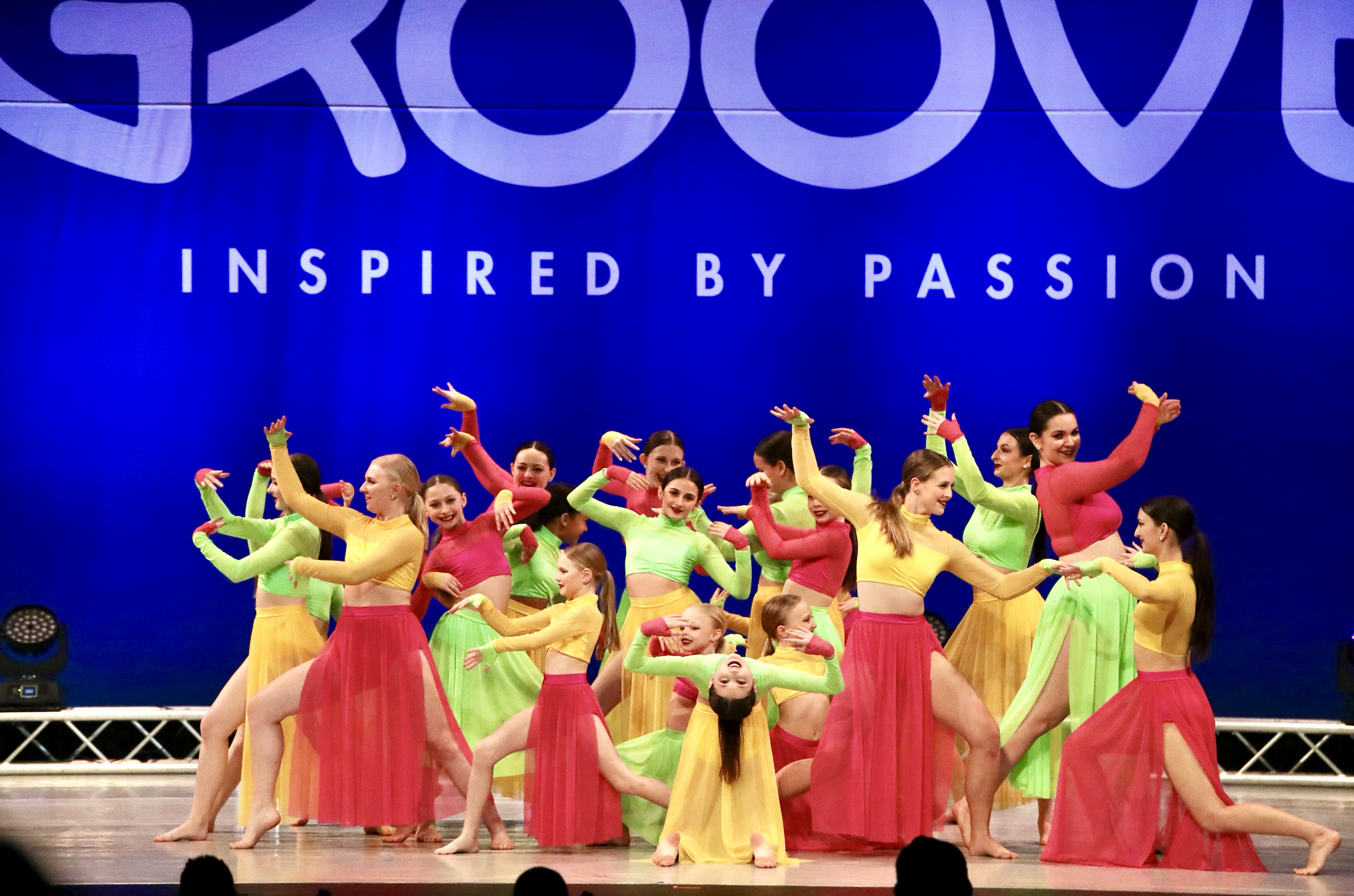Dance performance with multiple young girls in colorful costumes on stage at a competition, with a blue backdrop reading 'GROOVE' and 'Inspired by Passion.'