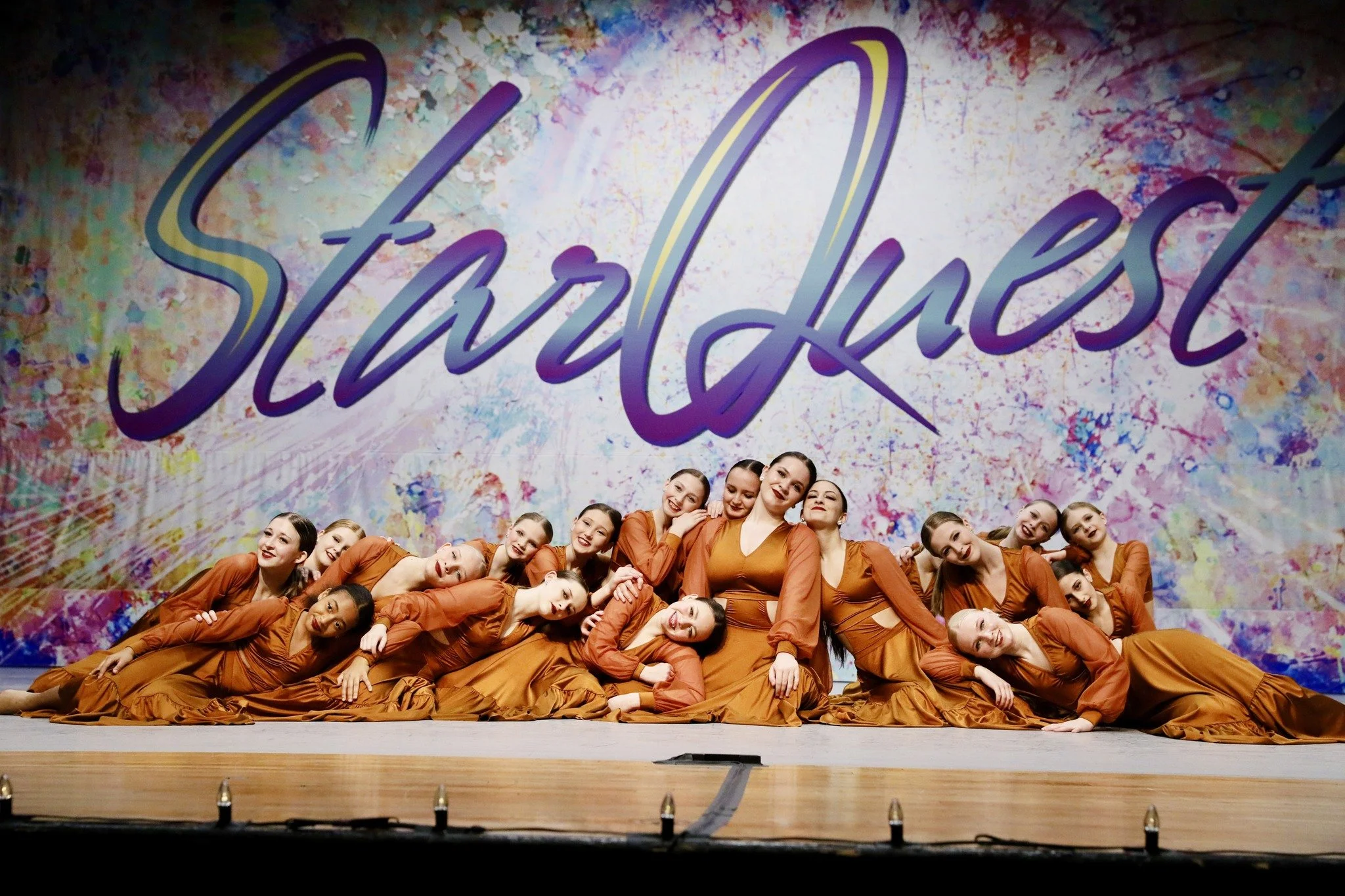 Group of young female dancers in matching orange costumes posing on stage in front of a colorful backdrop with the word 'Starquest' written in large cursive letters.