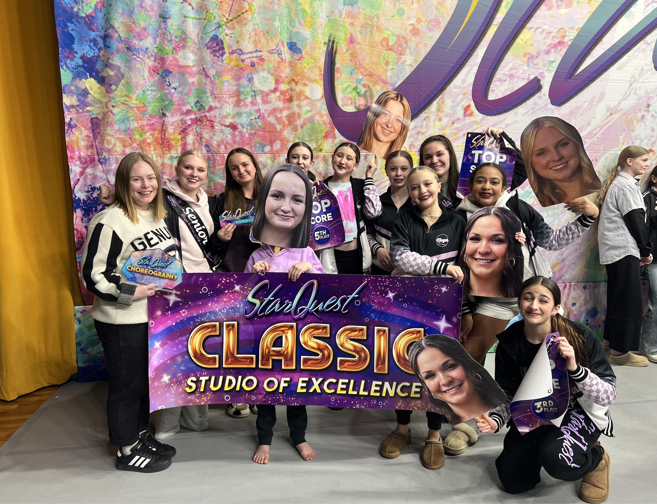 A group of young girls posing with a large banner that reads 'StarQuest Classic Studio of Excellence'. The girls are holding large cutouts of their faces and some are holding certificates, celebrating their achievement at a dance or performance compe
