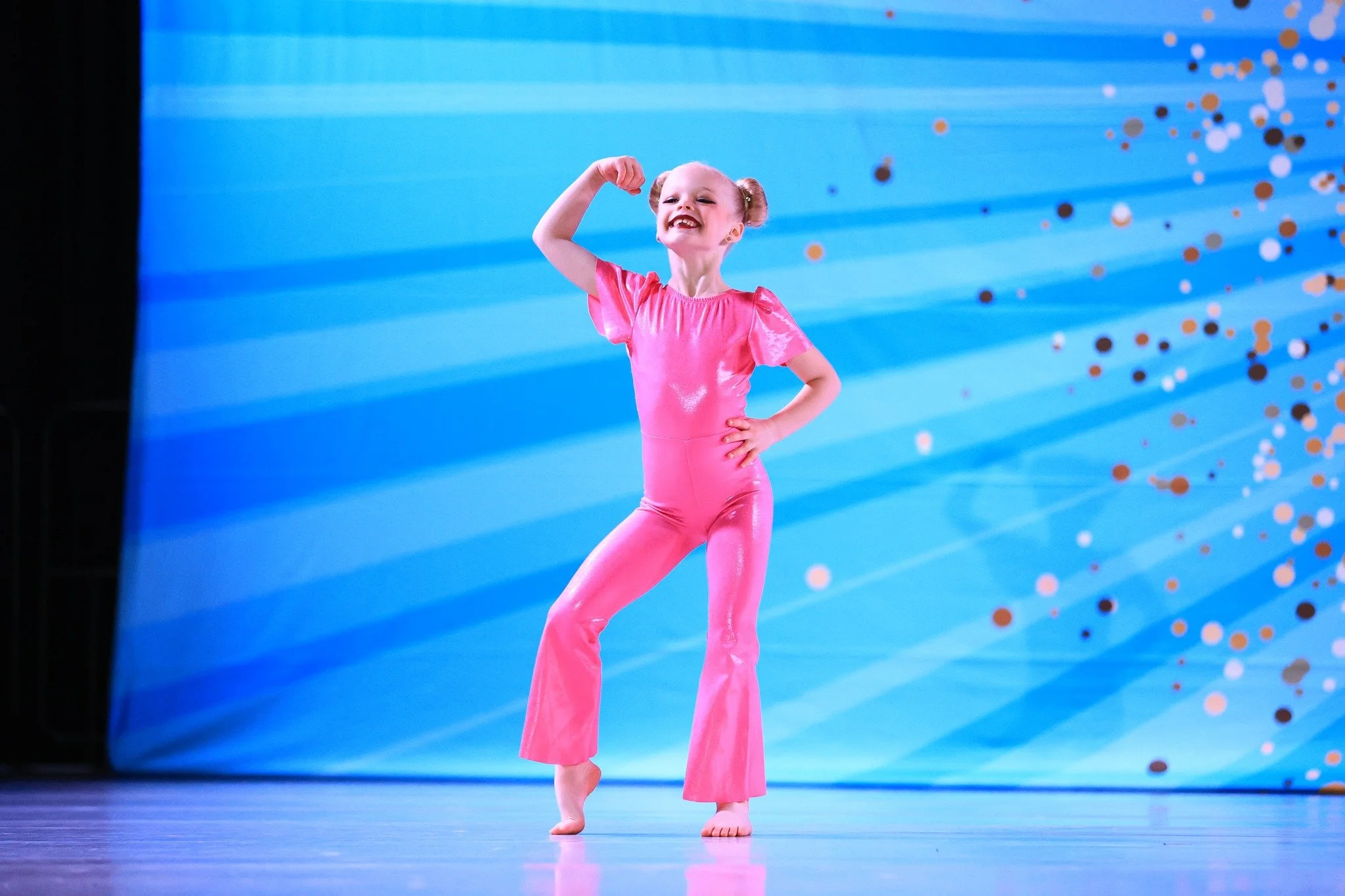 Young girl in a pink dance costume performing a pose on stage with a blue background and scattered confetti.