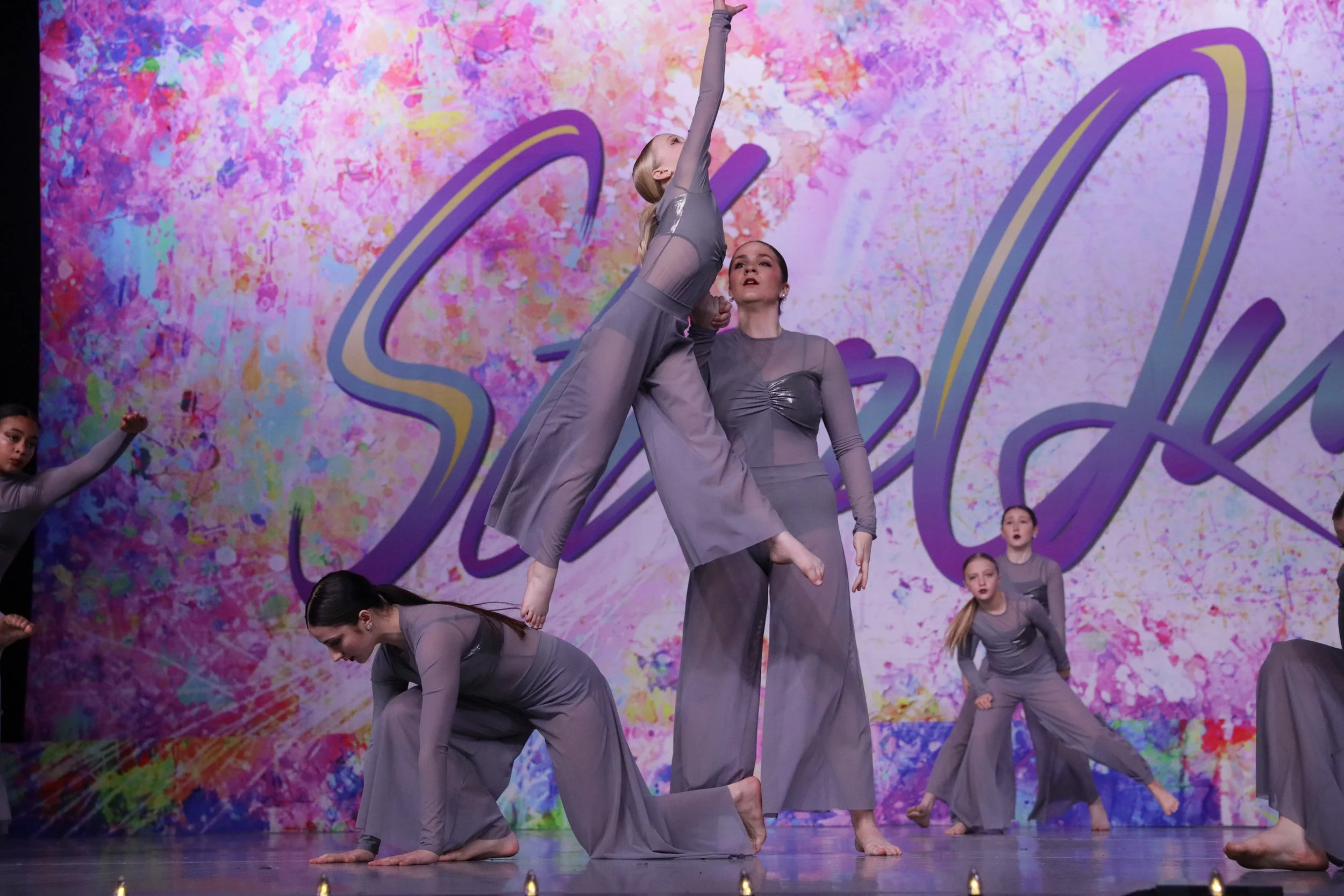 Group of young female dancers performing a contemporary dance on stage, dressed in gray costumes, with a colorful abstract background and the word 'Selah' written behind them.