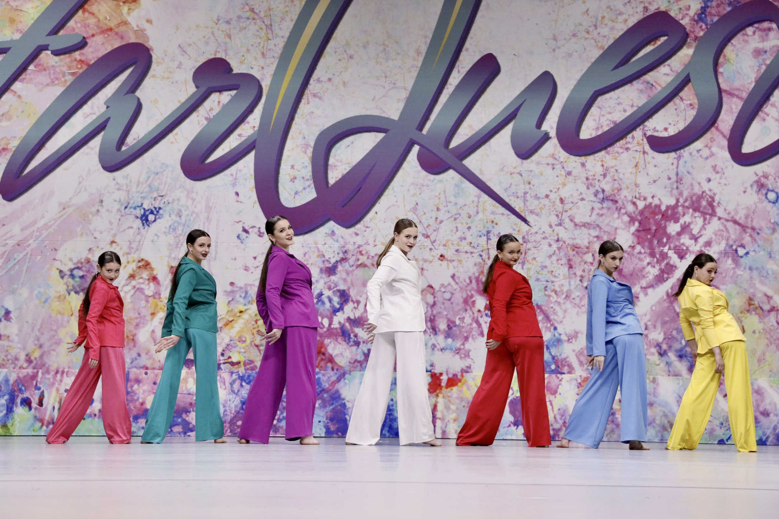 Group of seven young women in colorful dance costumes performing a dance on stage with a vibrant, splattered paint background that has the partial words 'wild' and 'sense' written on it.