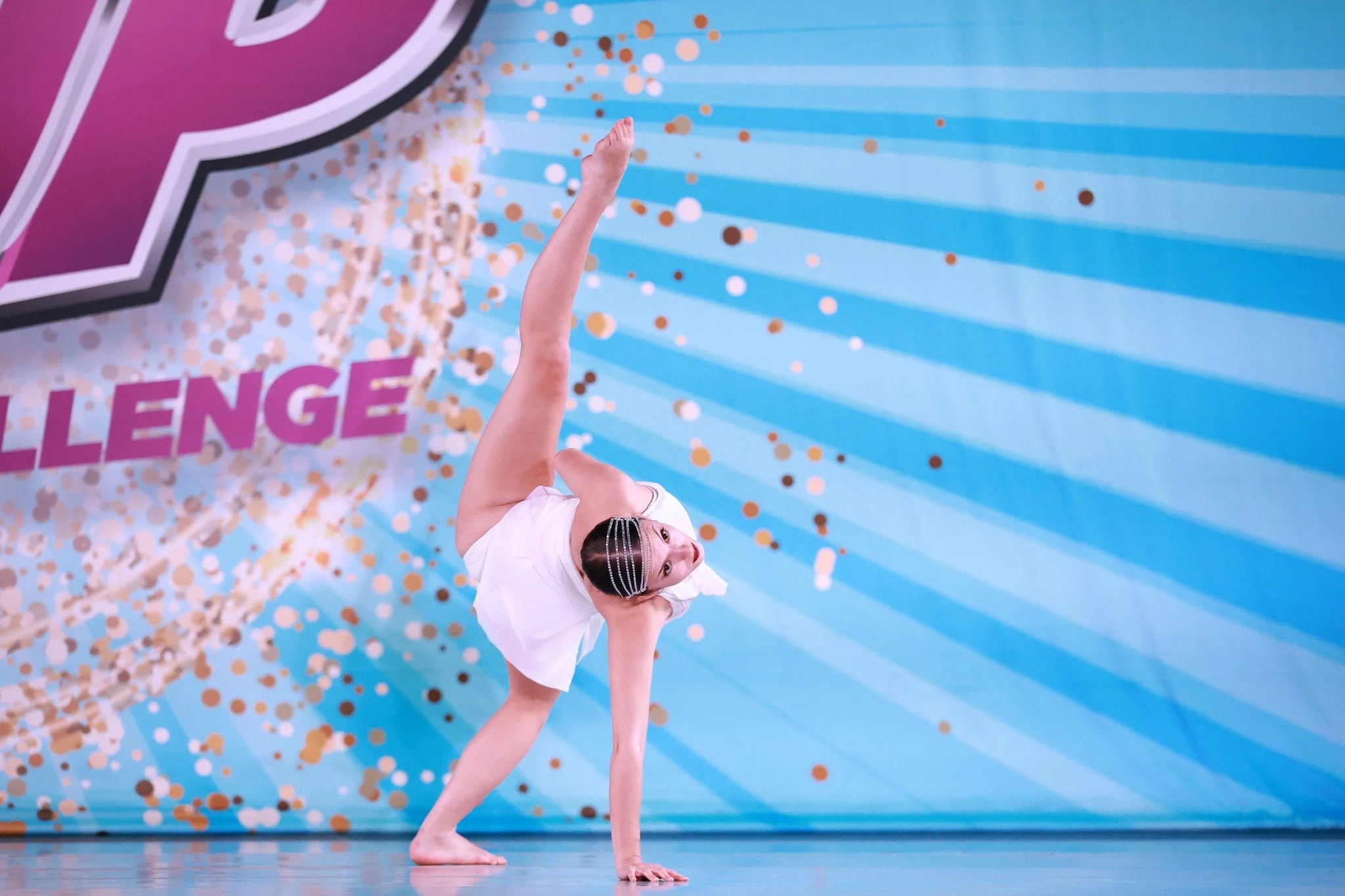 A young girl in a white dress performing a dance move on stage, balancing on her right hand with her left leg extended in the air, against a colorful backdrop with the words 'Dance Challenge' and confetti design.