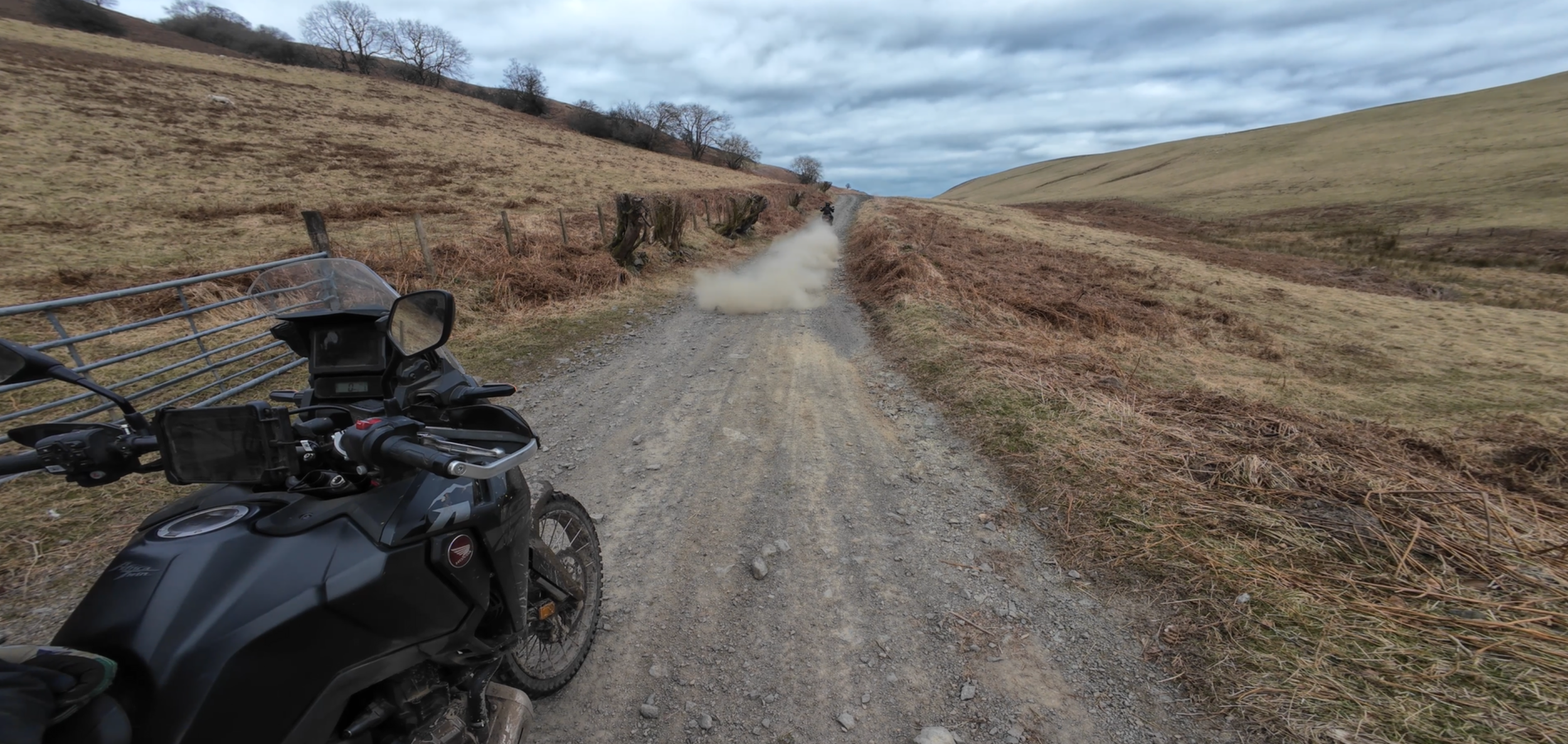 Motorcycle on a dirt road in a hilly landscape with dust cloud and cloudy sky.