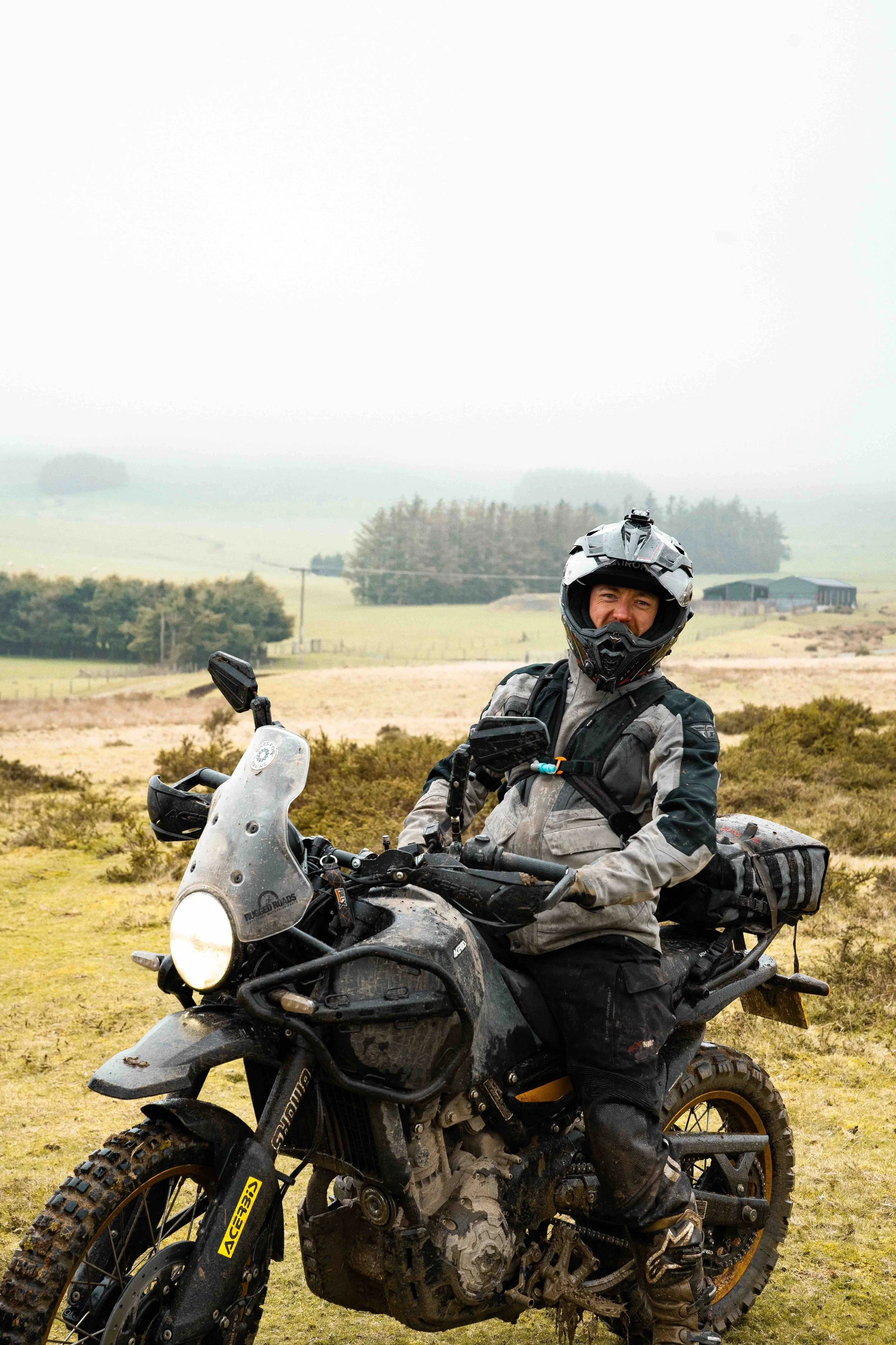 A person in motorcycle gear and a helmet sitting on a black adventure motorcycle in an open field with trees and a building in the background.