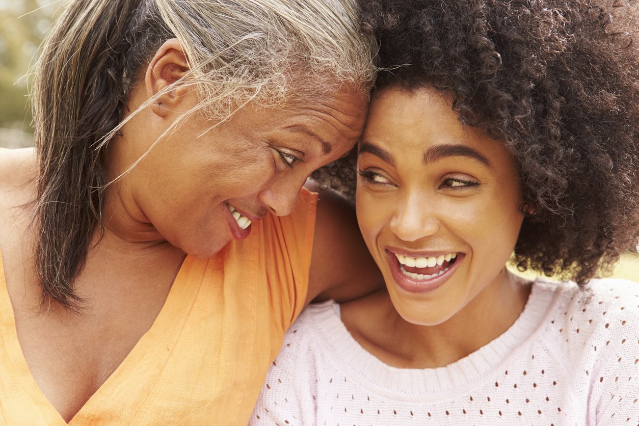 An older woman and a young woman are close together, smiling and touching foreheads in a joyful moment outdoors.