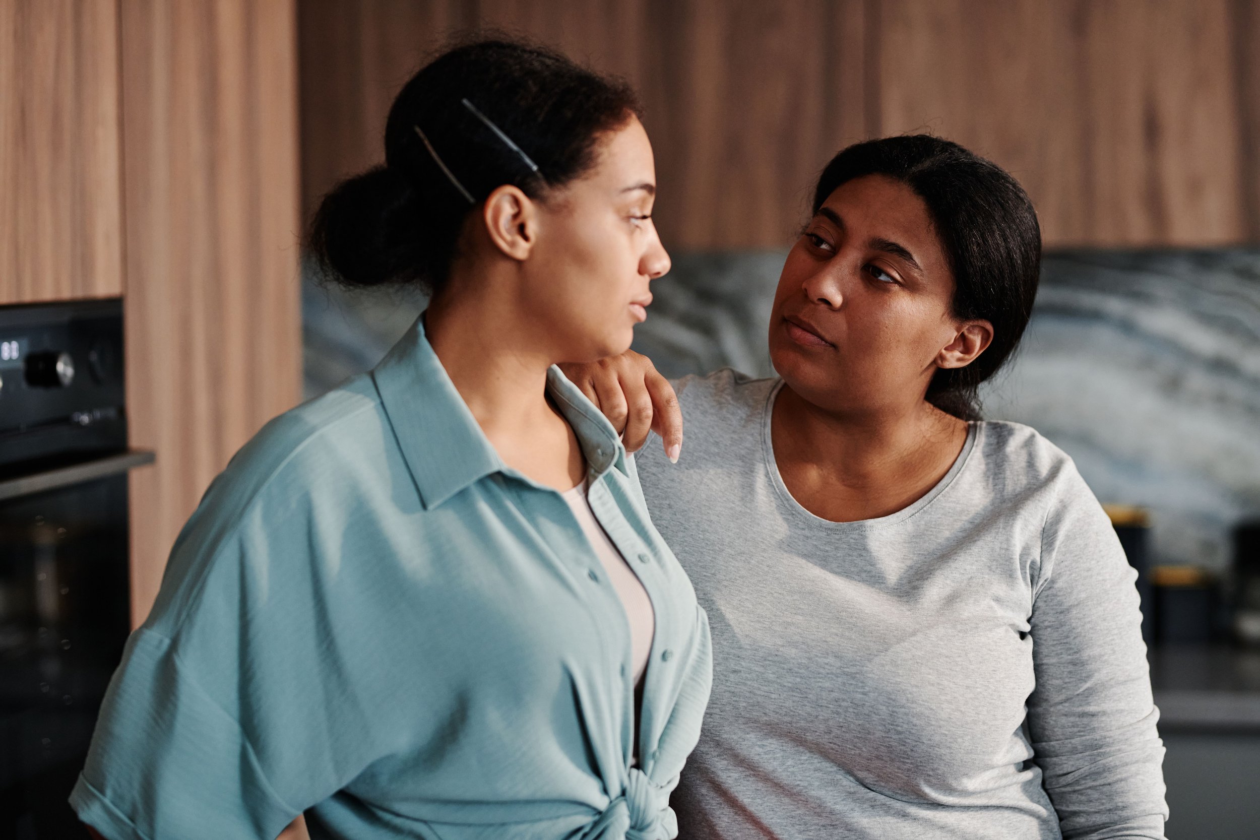 Two Black women, a mother and daughter, sitting together with a complex emotional expression, representing the love and difficulty that can coexist in mother-daughter relationships.