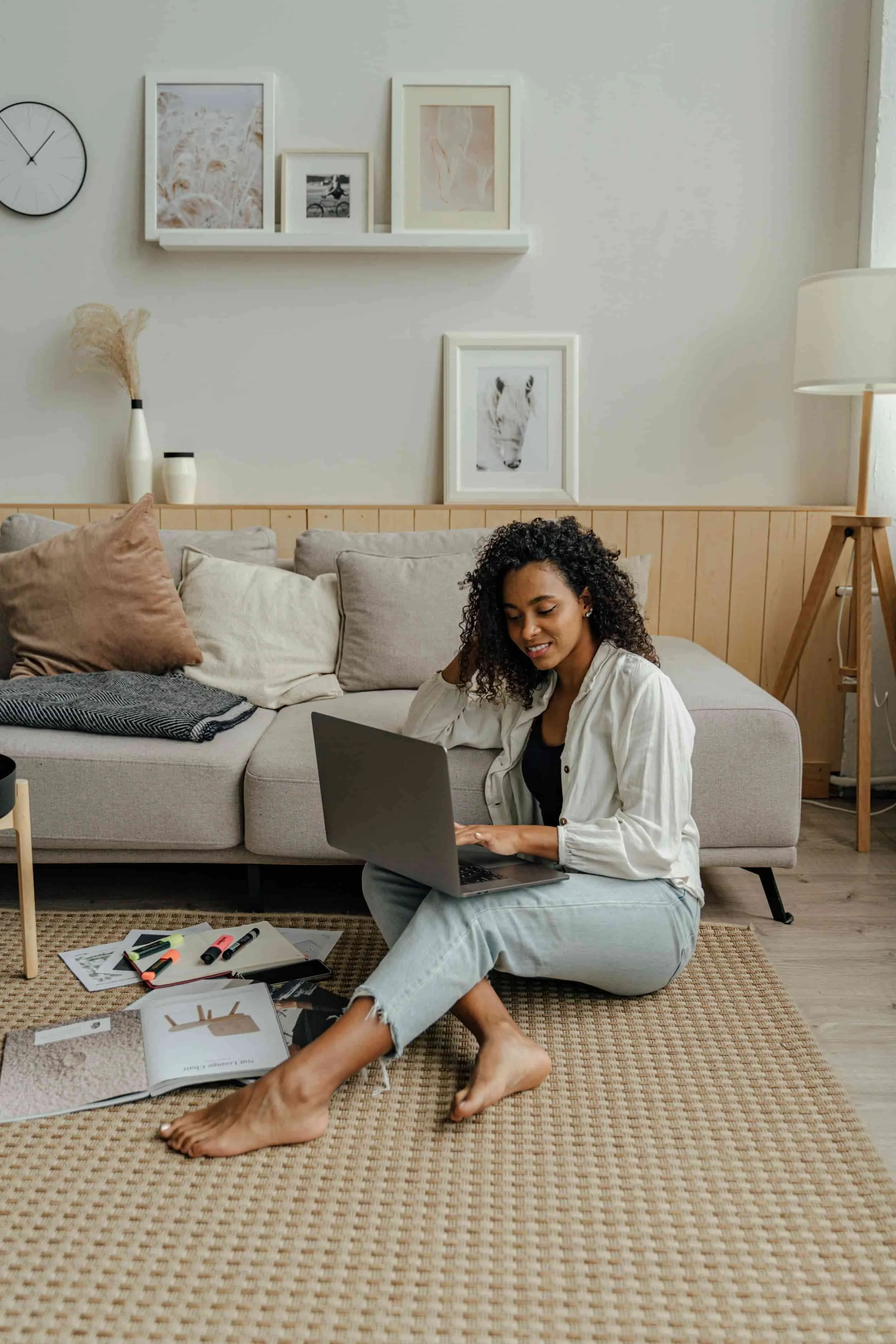 Woman sitting on the floor surrounded by books and pens smiling at her laptop
