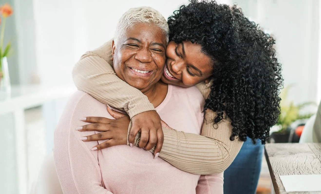 A woman hugging a smiling older man with white hair, in a bright, cozy room.