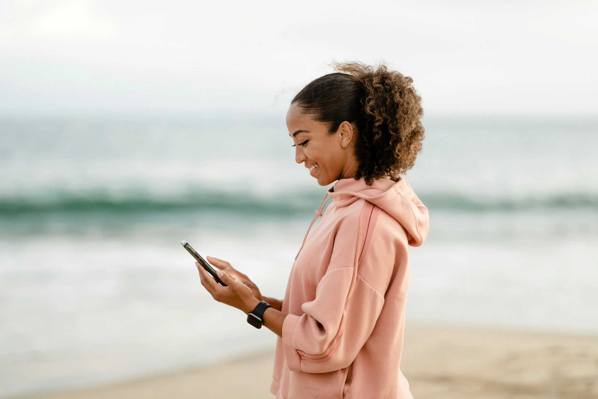 Woman walking on the beach smiling while looking at her phone