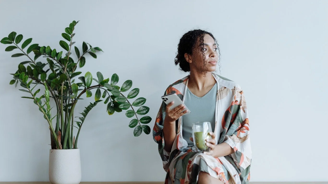 Woman sitting alone near a plant looking into the distance while holding her phone