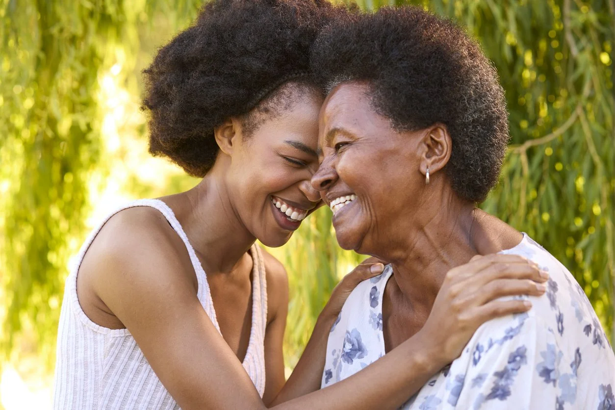 A young woman and an elderly woman smiling and touching foreheads outdoors with green foliage in the background.