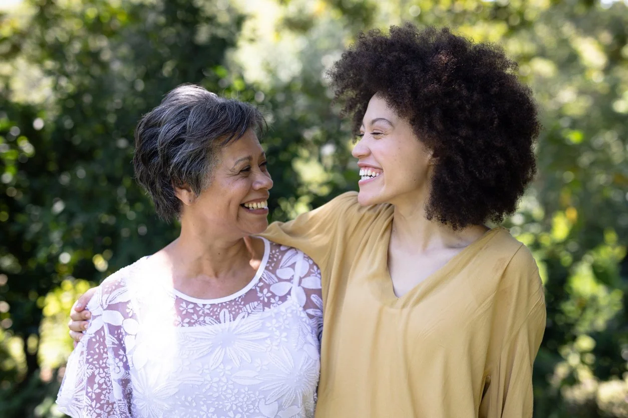 Two women smiling and embracing outdoors in a park with green trees in the background.
