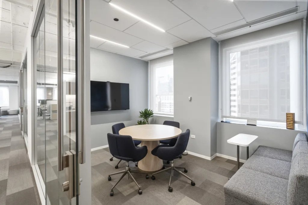 Modern office meeting room with a round table, five black swivel chairs, a wall-mounted TV, and a gray sofa near large windows with city views.