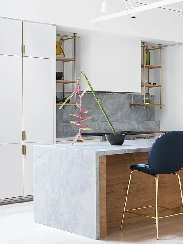 Modern kitchen with white cabinets, open wooden shelves, gray backsplash, a black bowl with pink and green decorative branches, and a blue chair with gold legs.