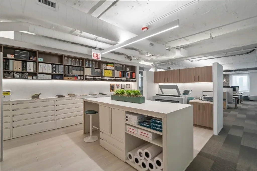 Modern office break room with white island and cabinets, shelves with binders, plants, printer, and workstations.