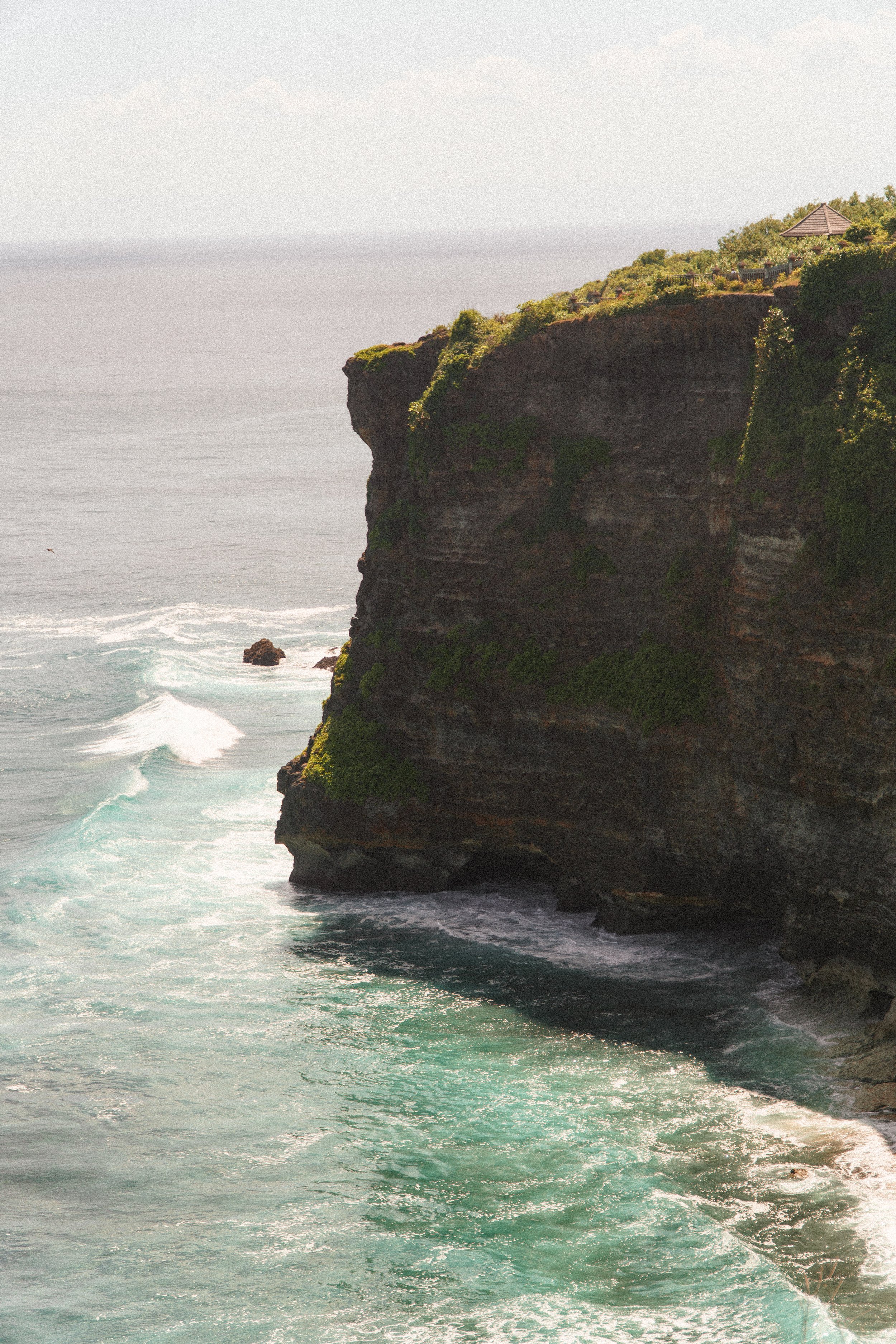 A coastal cliff with greenery and a small pavilion on top, overlooking the ocean with waves crashing below.