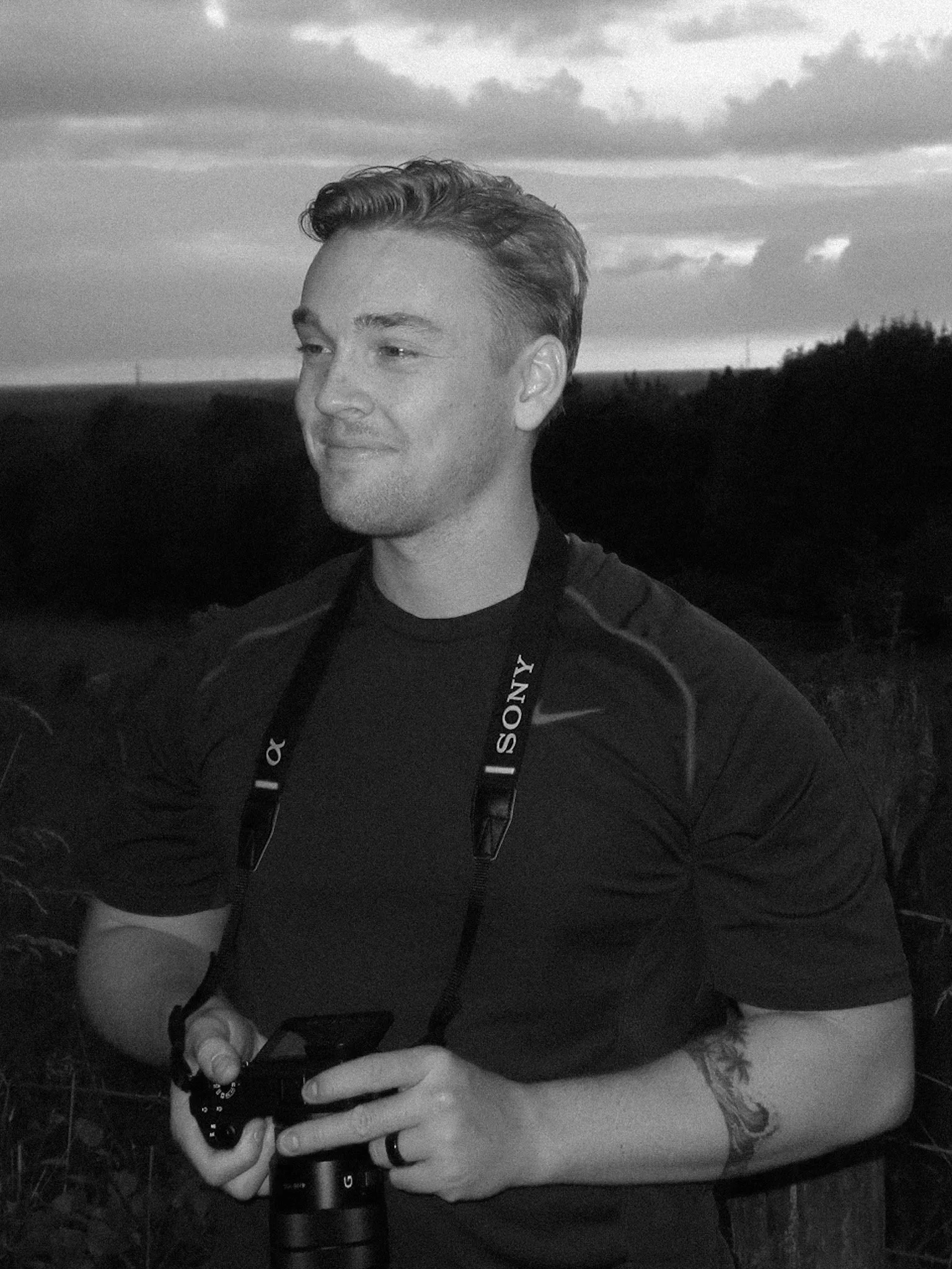 A young man with short, styled hair outdoors during sunset, holding a camera with a strap around his neck, and smiling slightly while looking off to the side.