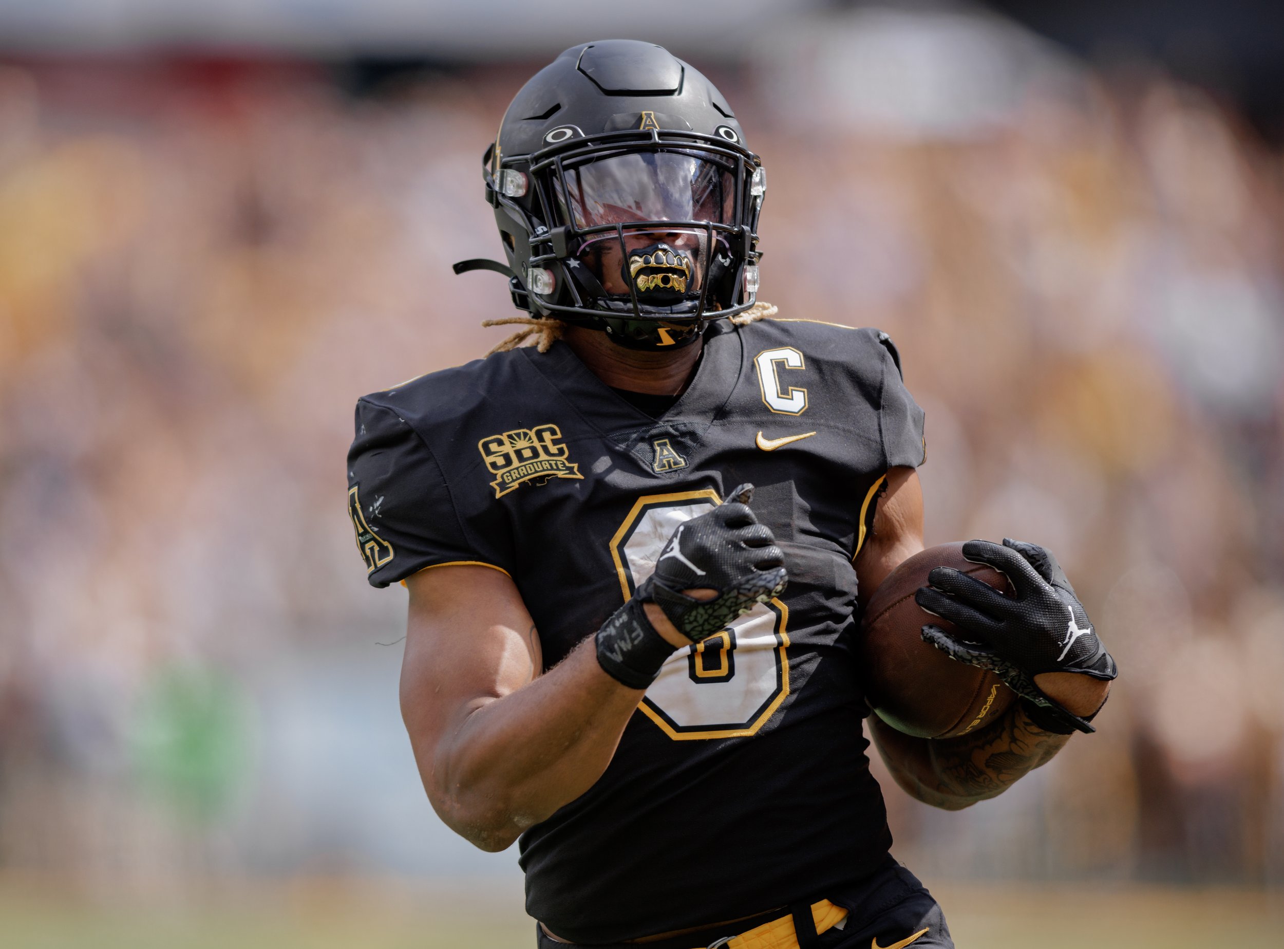 A football player in a black uniform with gold accents, wearing a helmet, running with a football in hand.