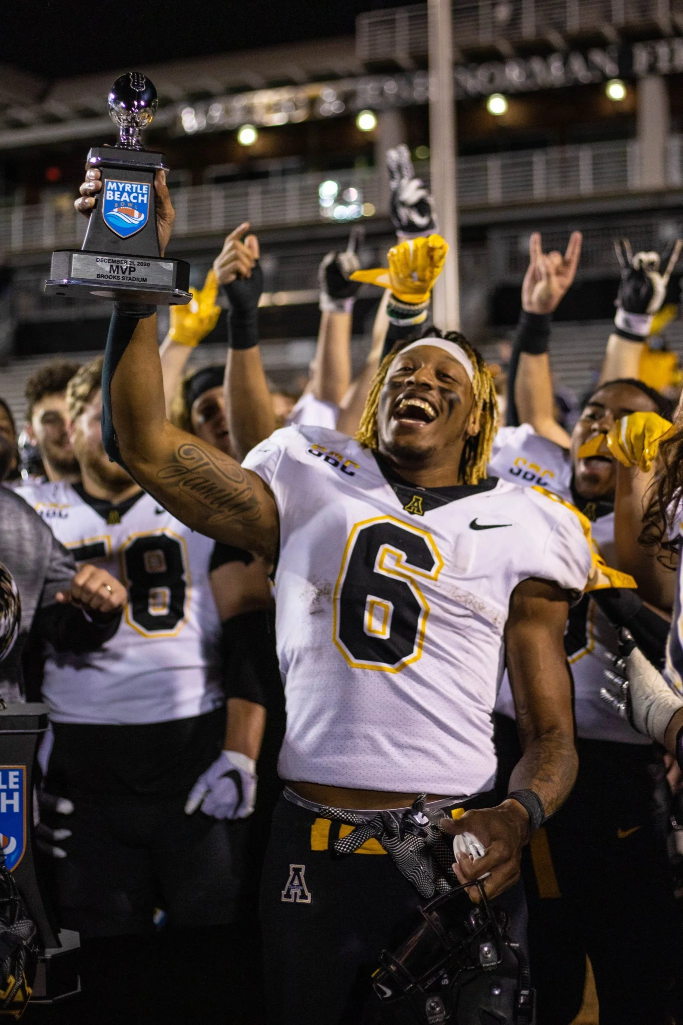 A football player in a white jersey with the number 6 and an 'A' logo celebrates with a trophy after winning a game, surrounded by teammates on a football field.