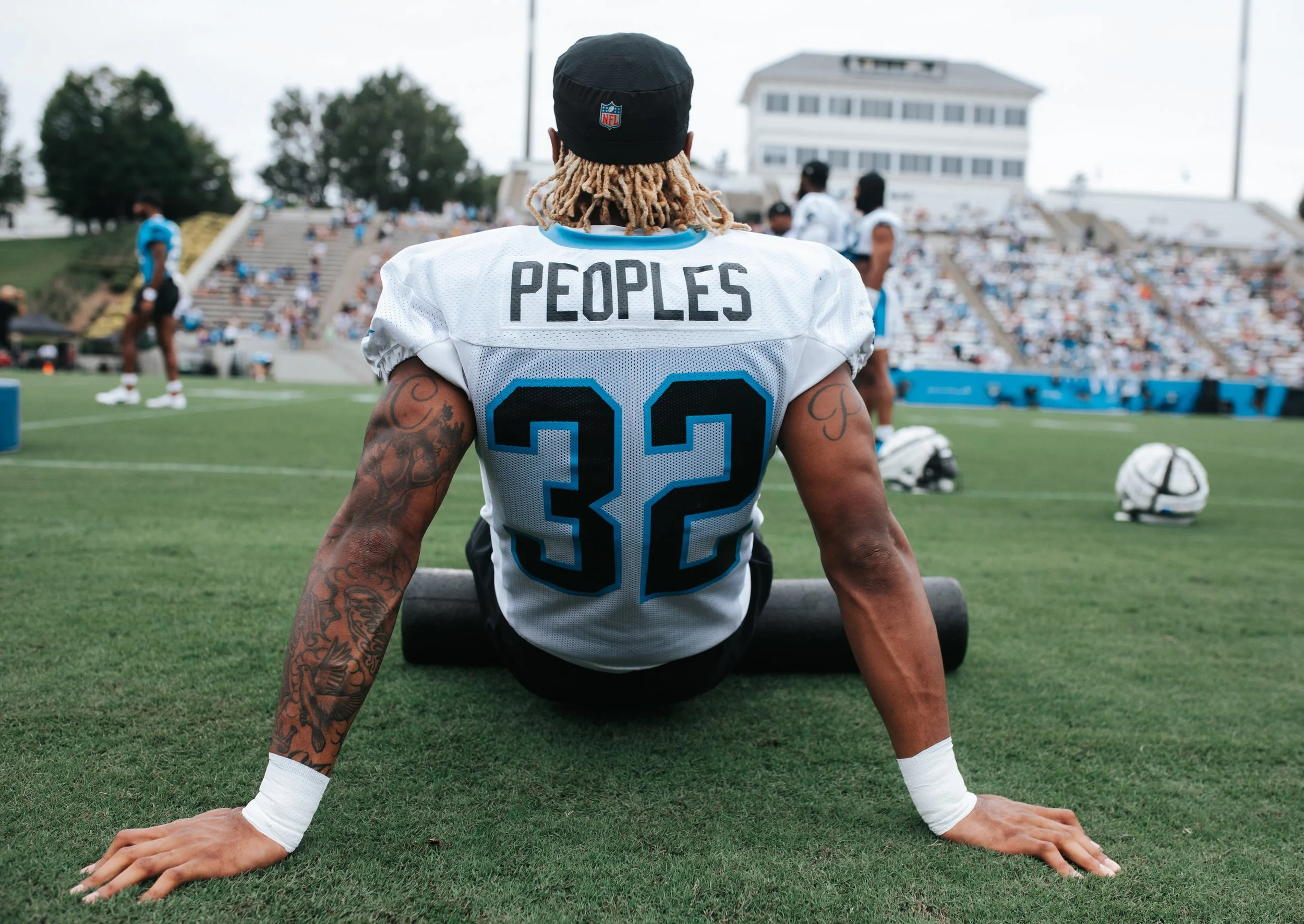 American football player sitting on the sideline of a football field, wearing a white jersey with the name 'PEOPLES' and number '32' on the back, and a black cap. The player has tattoos on arms and long hair, and is surrounded by teammates and equipment, with stands filled with spectators in the background.