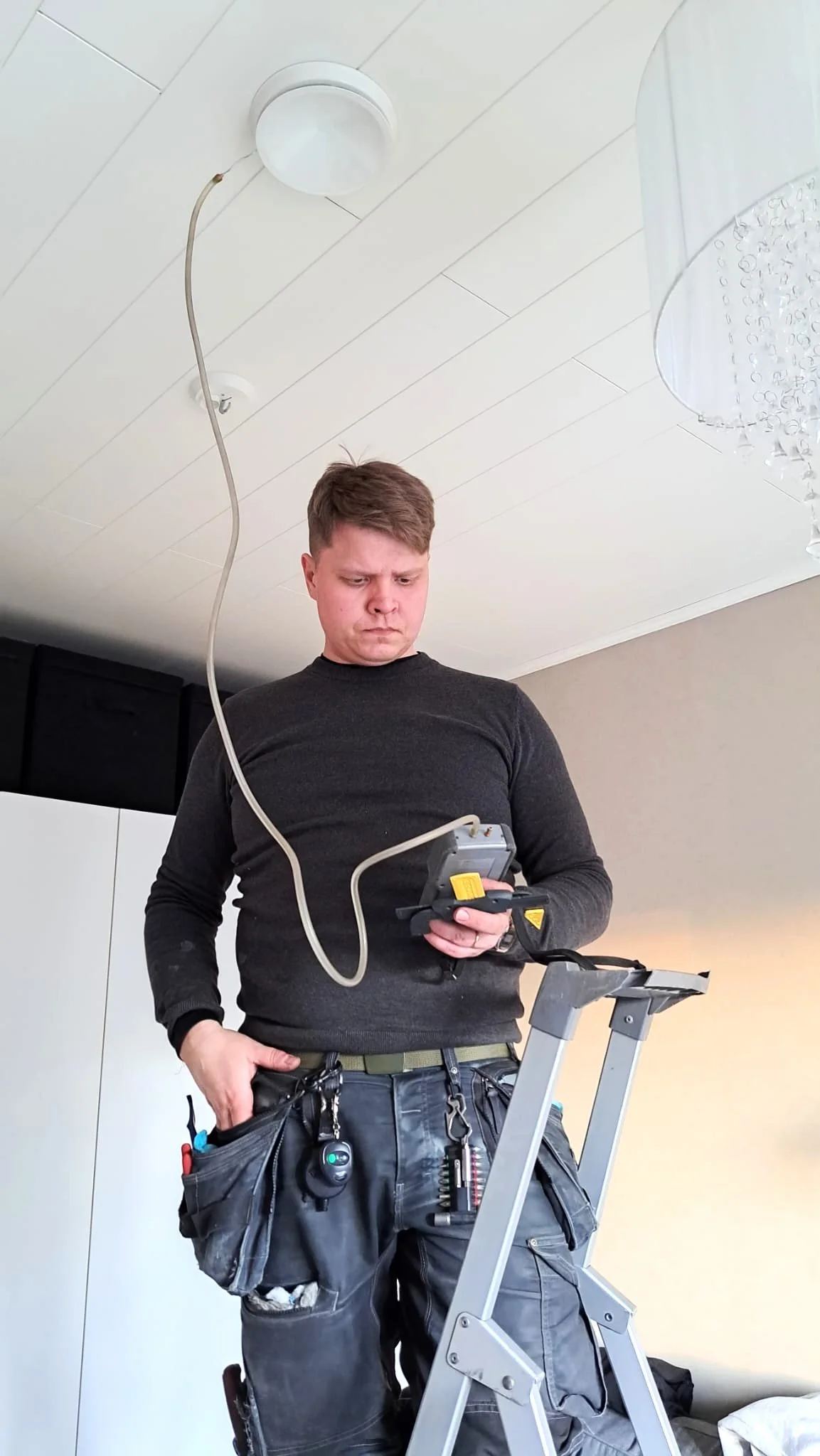Ilmateknikko Antti Lepikko tarkistaa mittarista ilmamäärien virtauksen työvaatteissaan A-tikkailla.