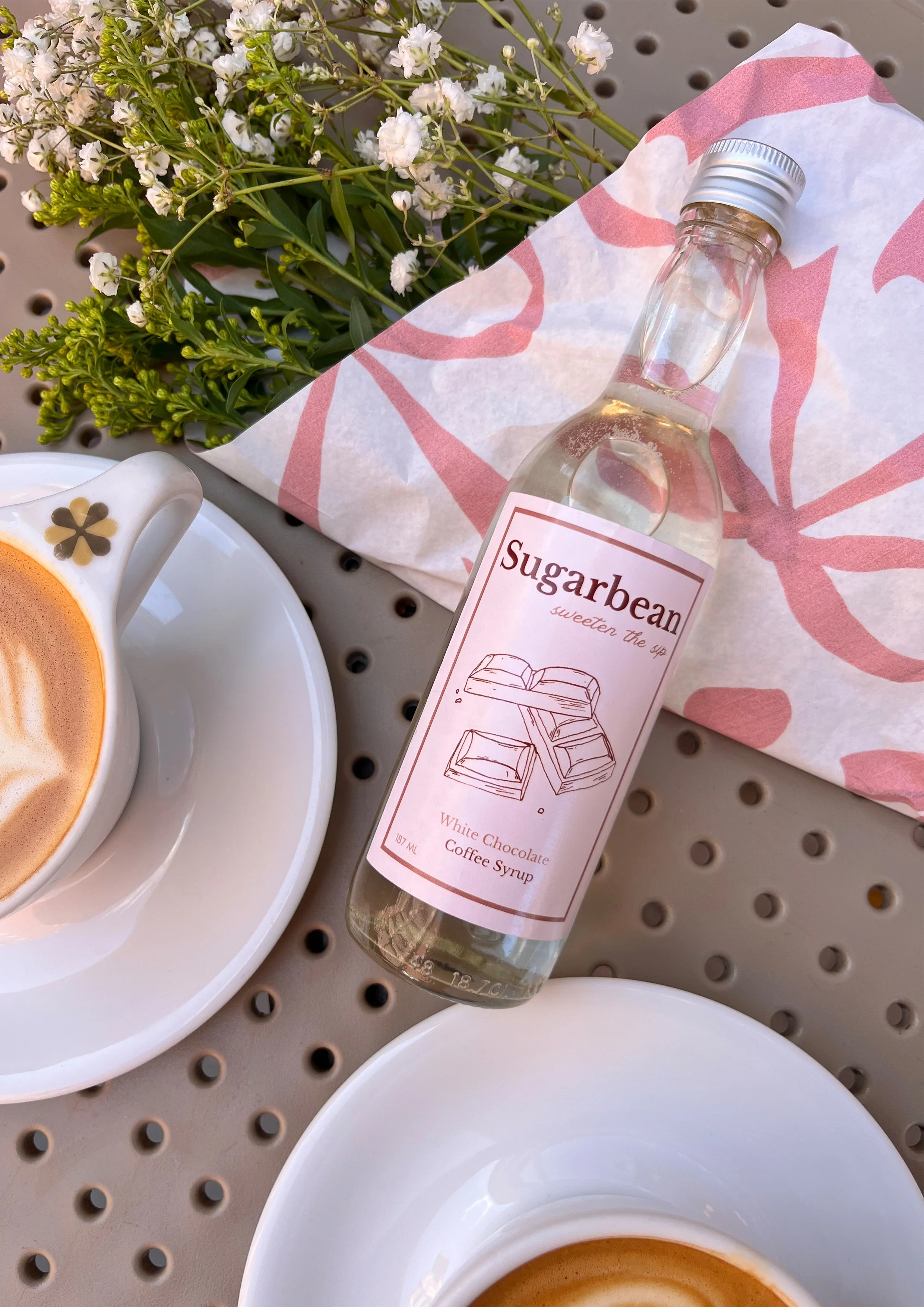 a white ceramic cup filled with coffee on a white saucer, a tall glass bottle of Sugarbeand white chocolate coffee syrup, a bouquet of white and green flowers wrapped in pink and white paper, and a gray perforated table surface.