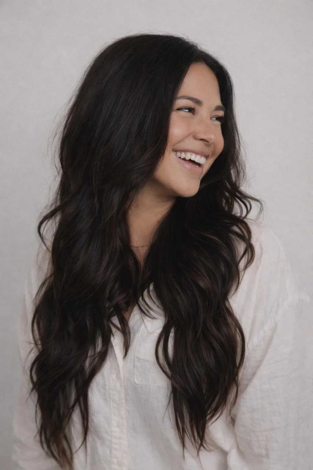 A woman with long, dark, wavy hair smiling and looking to her right, wearing a white shirt.