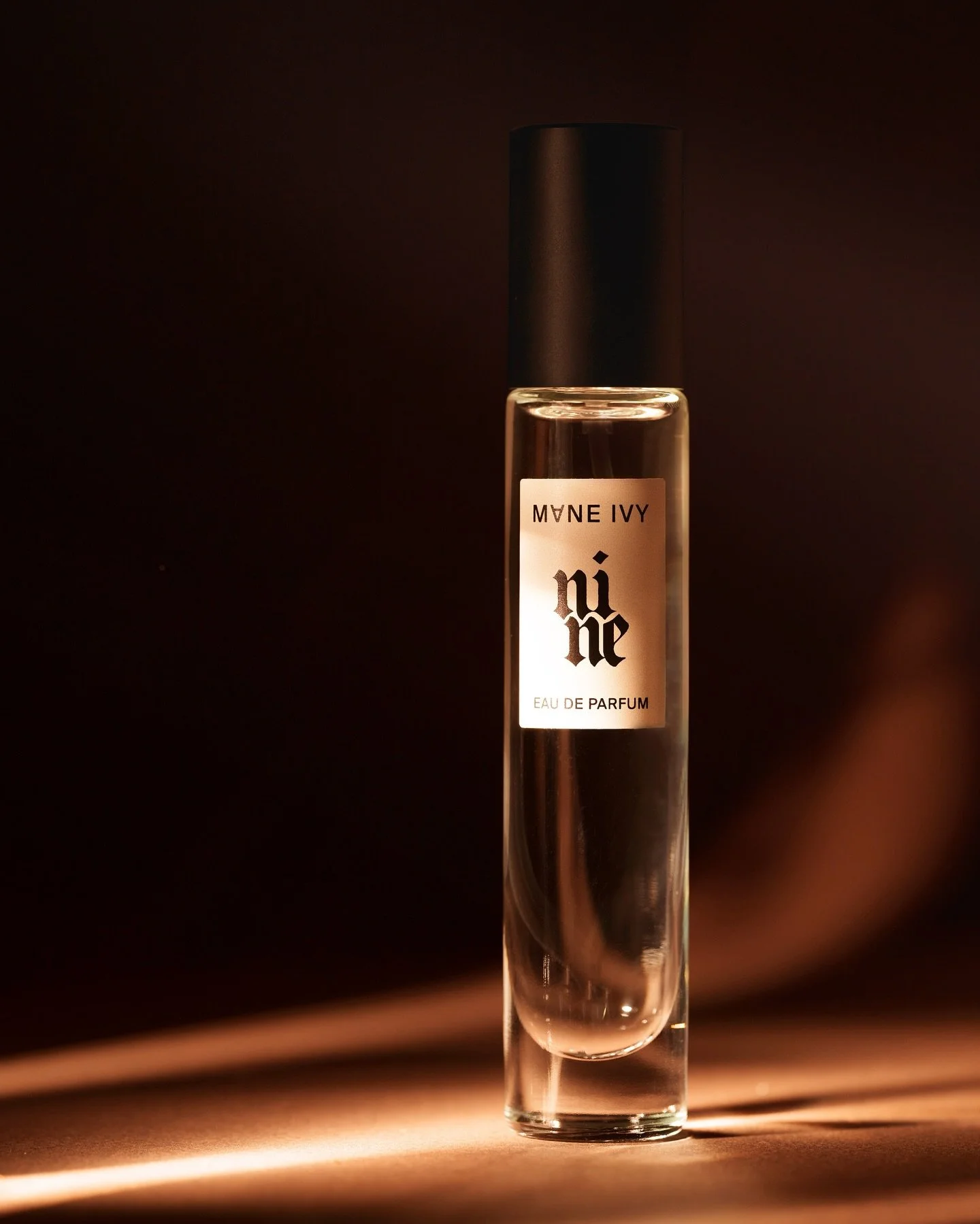 A tall, slender glass perfume bottle with a black cap, labeled 'MNNE IVY' and 'nina' in gothic font, set against a dark background with soft lighting.