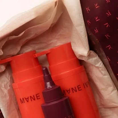 Two cylindrical orange containers with black text, partially wrapped in tissue paper, next to a dark purple lipstick or lip gloss tube with a clear cap.