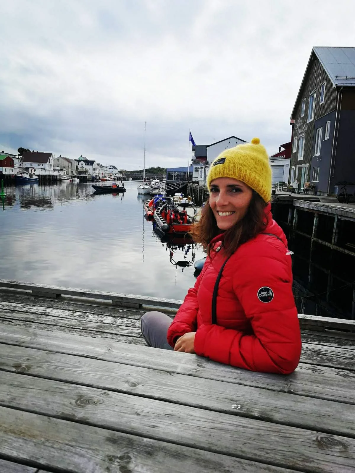 Una donna sorridente indossa un berretto giallo e una giacca rossa, seduta su un molo di legno vicino all'acqua, con case e barche in un paesaggio marittimo in una giornata nuvolosa.