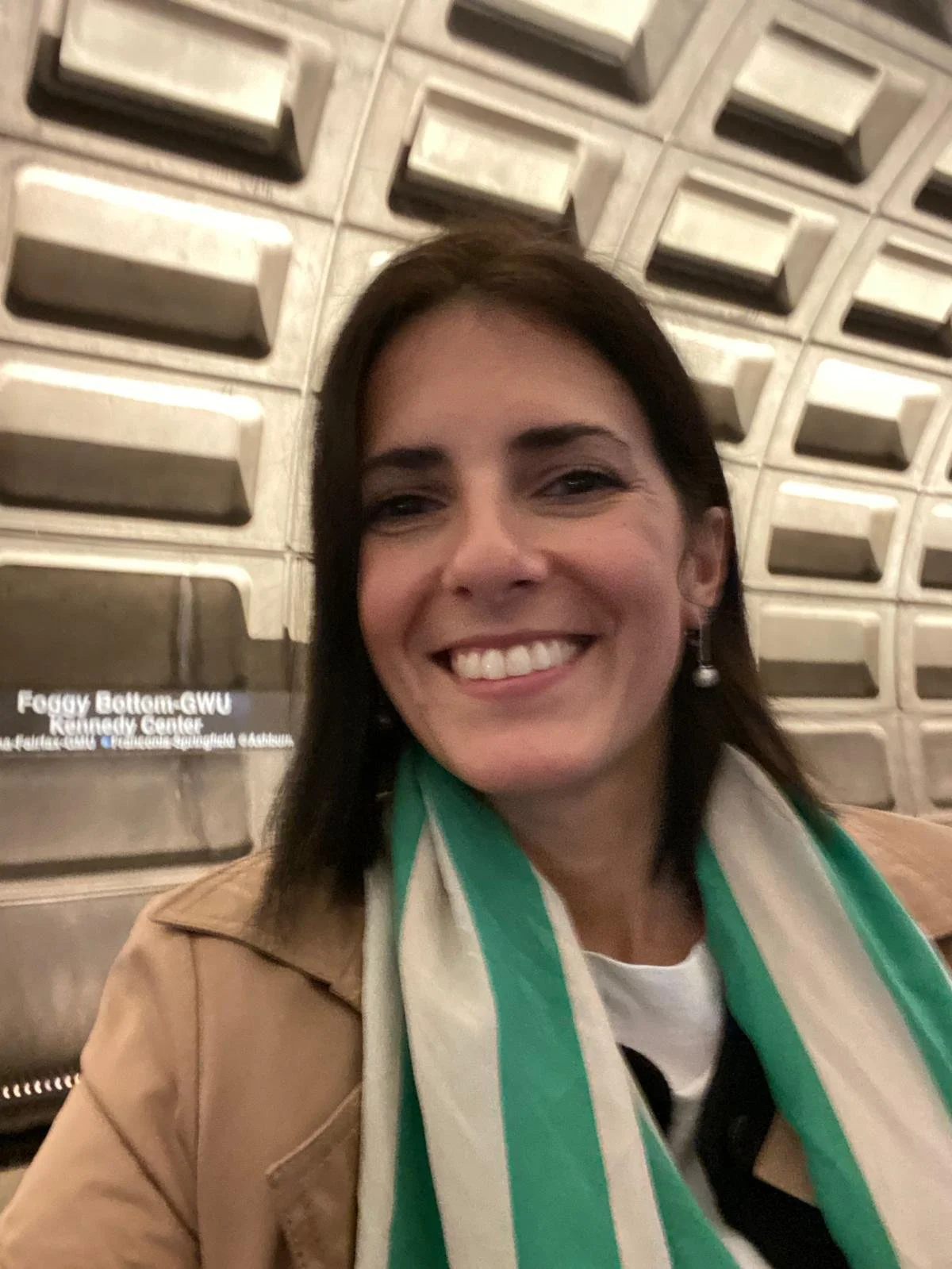 Una donna sorridente con capelli castani, indossa una sciarpa verde e bianca e una giacca marrone, seduta davanti a una parete di blocchi metallici.