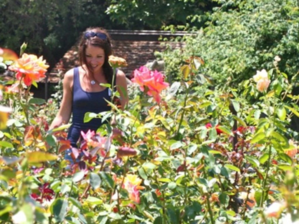 A woman in a blue top with sunglasses on her head smiling and walking through a garden filled with pink, orange, and white flowers with green leaves.