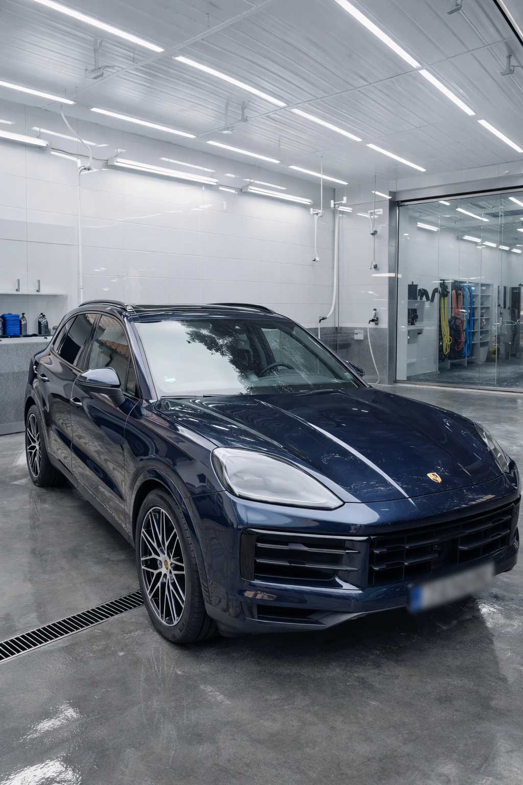A dark blue Porsche SUV parked inside a well-lit, clean garage with white walls and ceiling, and transparent partitions showing car cleaning equipment and tools.