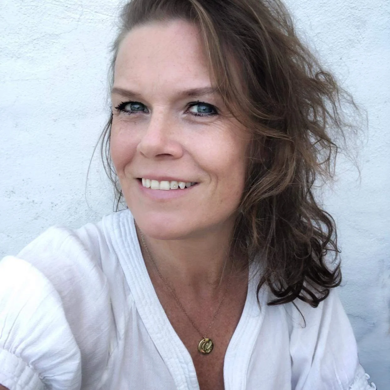 Close-up of a woman with wavy brown hair, blue eyes, smiling, wearing a white top and a gold necklace with a circular pendant, against a plain white wall background.