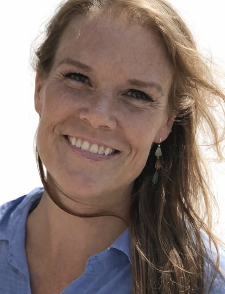 Close-up photo of a woman with red hair, freckles, and blue eyes, smiling and wearing a blue shirt and earrings.