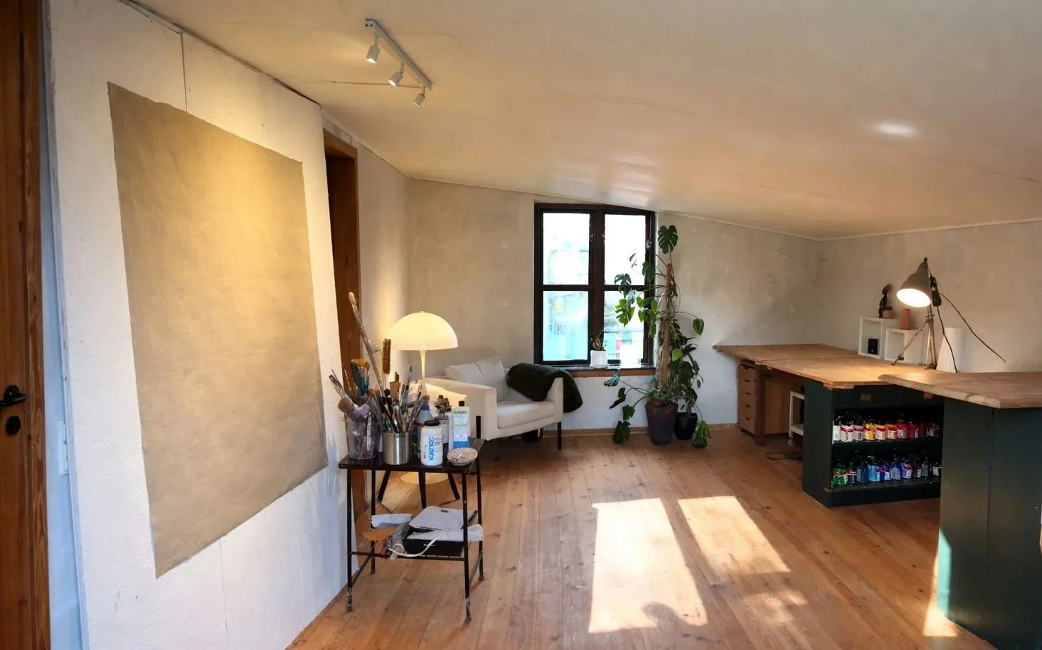 Cozy room with a white sofa next to a window, a potted plant, a wooden table with art supplies, and a bar counter with snacks, illuminated by a desk lamp and ceiling lights.