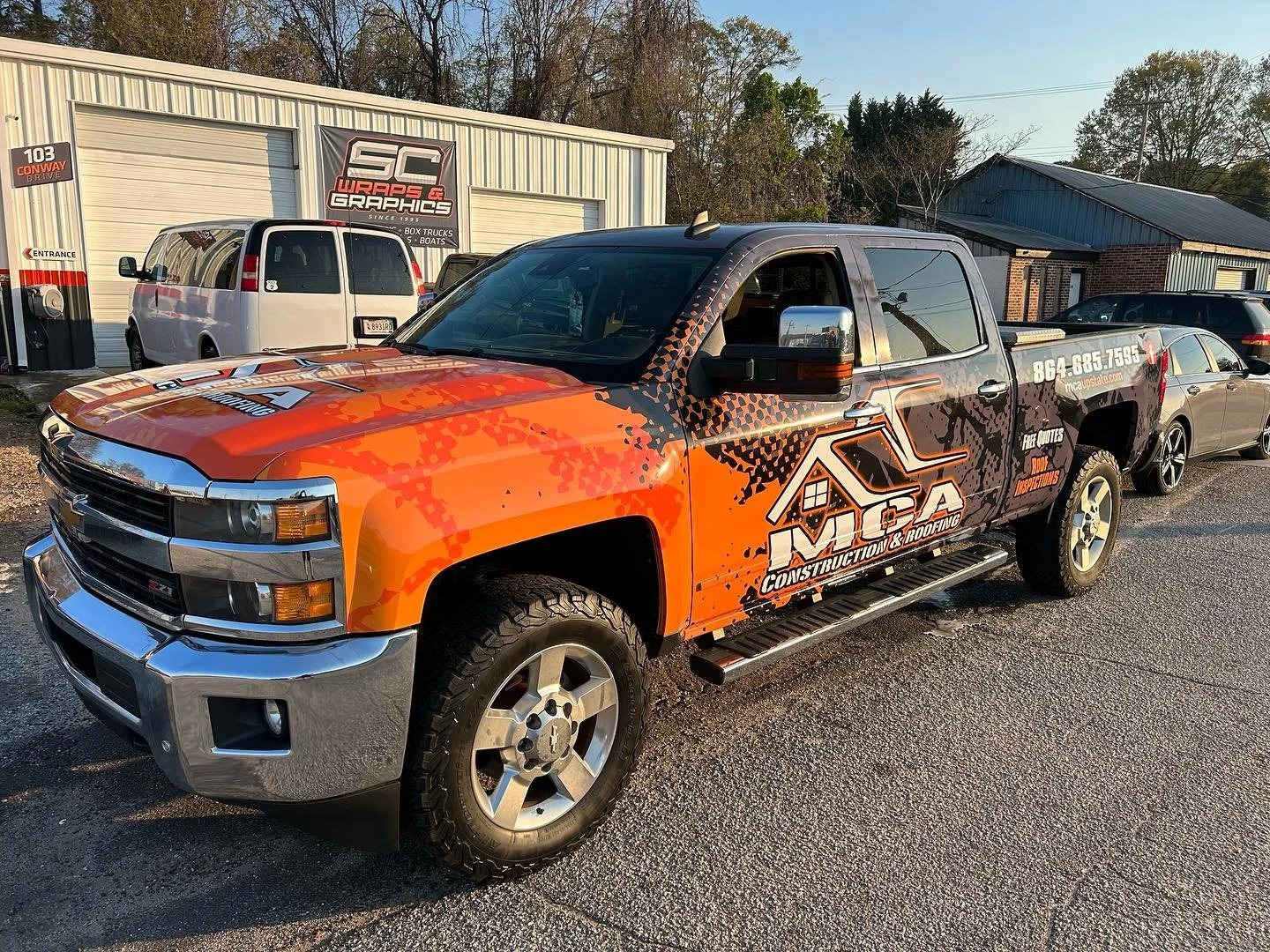 A pickup truck with a custom orange and black camouflage wrap advertising 'MCA Construction & Roofing,' parked outside a building with a sign for 'WS Graphics.' Multiple vehicles are visible around the truck, including a white van, a gray car, and ot