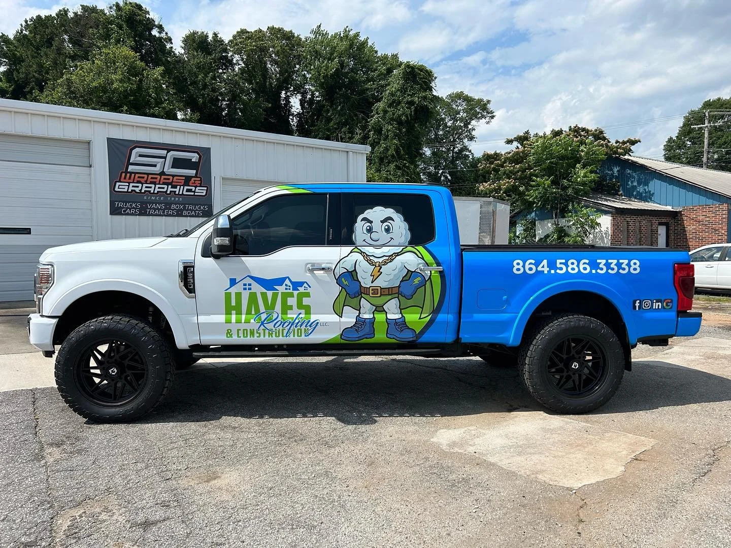 A pickup truck for Haves Roofing & Construction parked outdoors. The truck has large black tires, a white cab, and a blue bed featuring a cartoon superhero character with a muscular build, cape, and lightning bolt emblem. The truck includes contact i