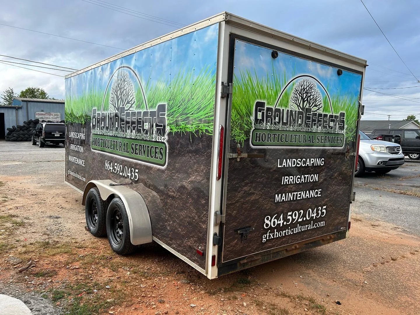 Mobile trailer advertising Ground Effects horticultural services with a logo of a leafless tree inside a circle, green grass illustration, and text listing services: landscaping, irrigation, maintenance, with phone number and website.