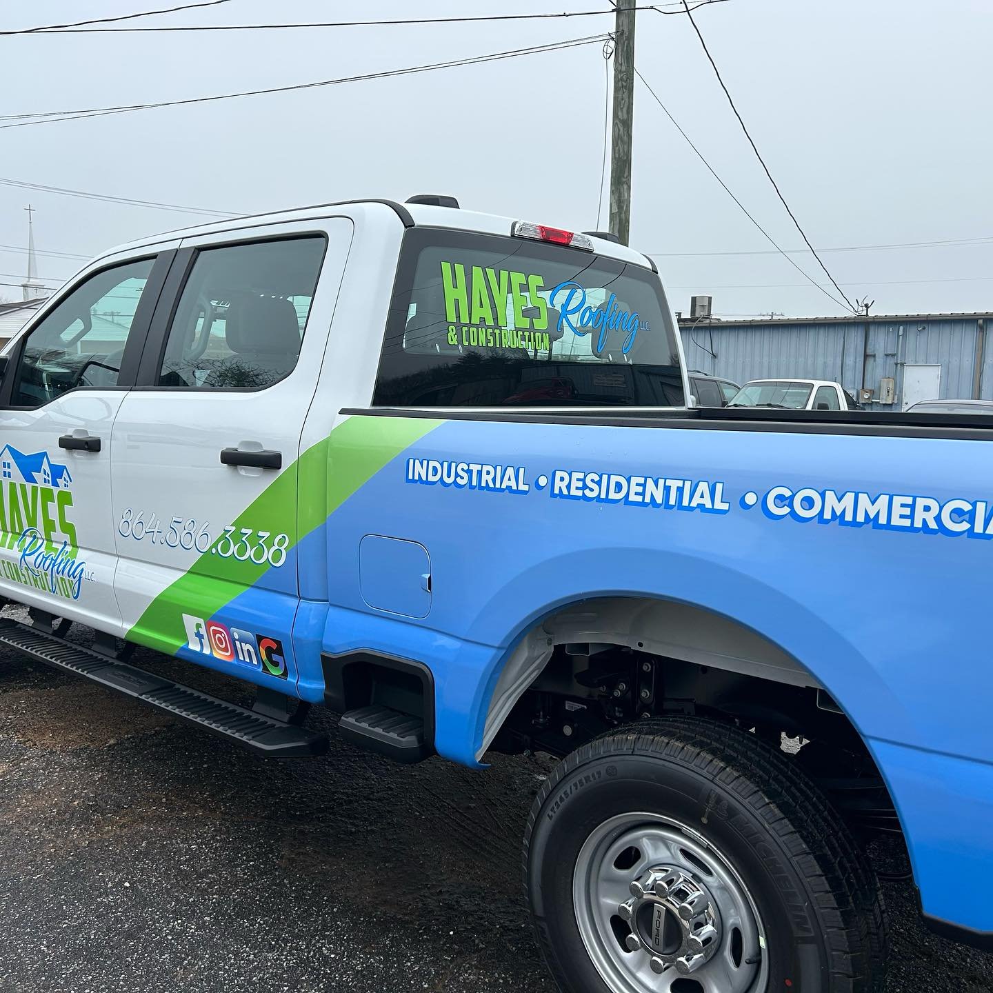 A pickup truck with promotional graphics for Hayes Roofing & Construction, displaying contact number, social media icons, and a list of services including industrial, residential, and commercial roofing, parked on a gravel lot under a cloudy sky.