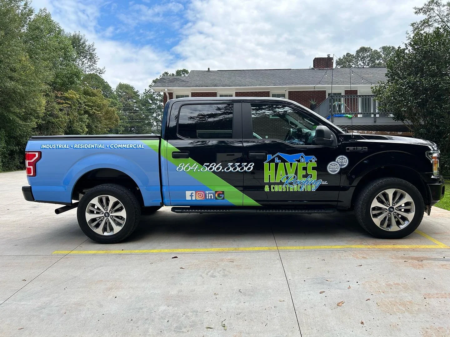 Side view of a black and blue pickup truck with green and blue branding for 'Haves Roofing & Construction LLC,' parked on a driveway with house and trees in the background.